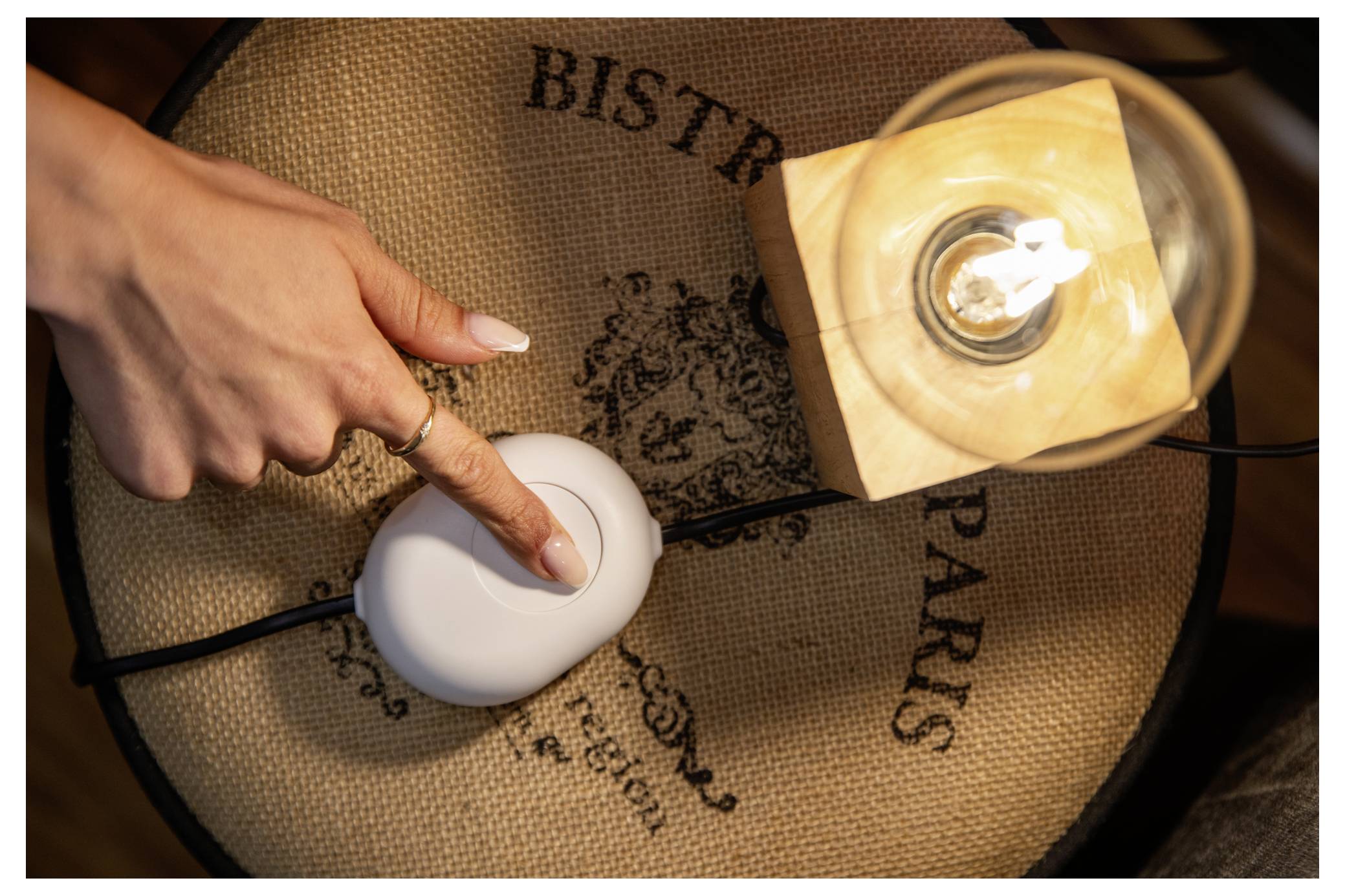 A hand with a ring points at a white light switch on a table next to a glowing lamp, set on a rustic surface.