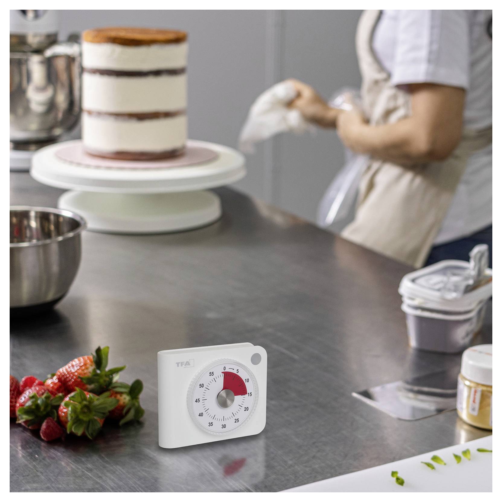 Une personne décore un gâteau sur un plateau tournant dans une cuisine, avec des fraises sur le plan de travail et un minuteur affichant 10 minutes restantes.