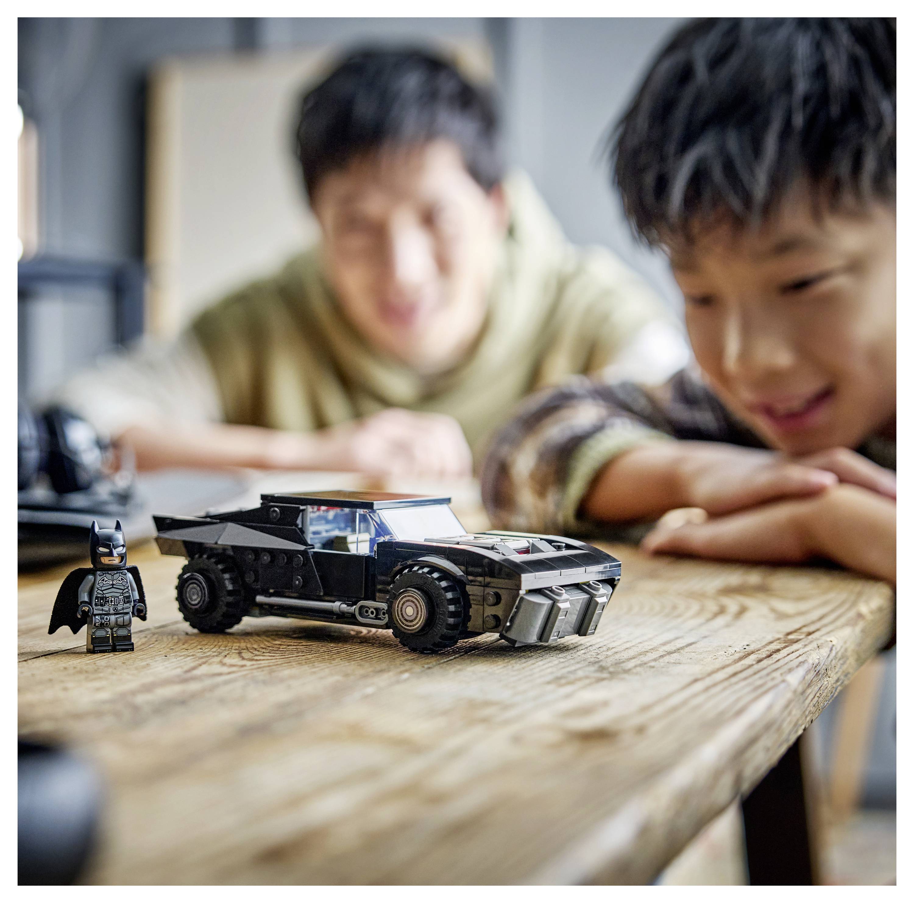 Un enfant souriant à une voiture jouet ressemblant à la Batmobile sur une table en bois, avec un adulte en arrière-plan qui regarde avec bonheur.