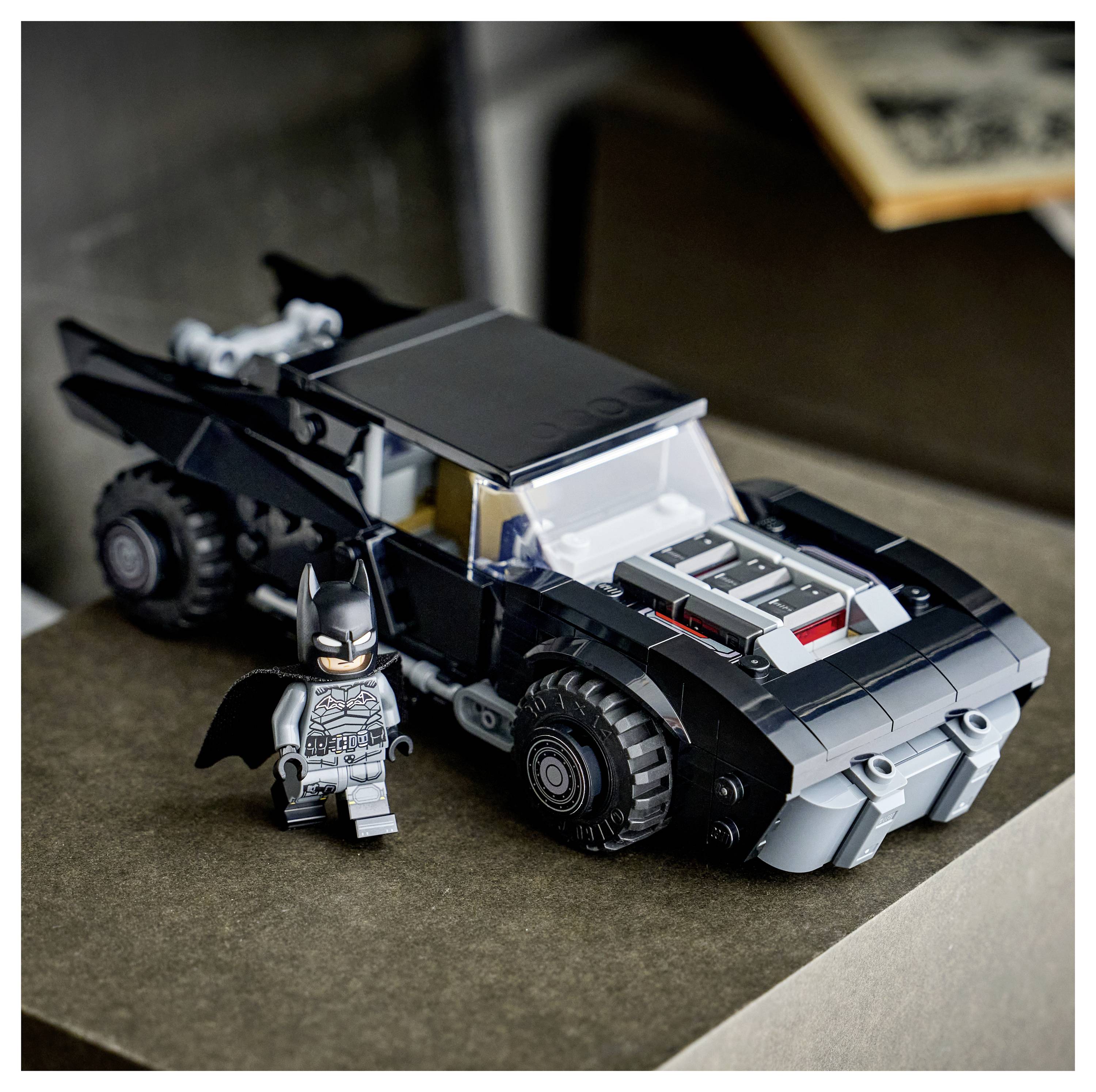 Une petite voiture jouet noire avec des détails argentés est stationnée sur une surface, accompagnée d'une petite figurine jouet vêtue en superhéros en noir.