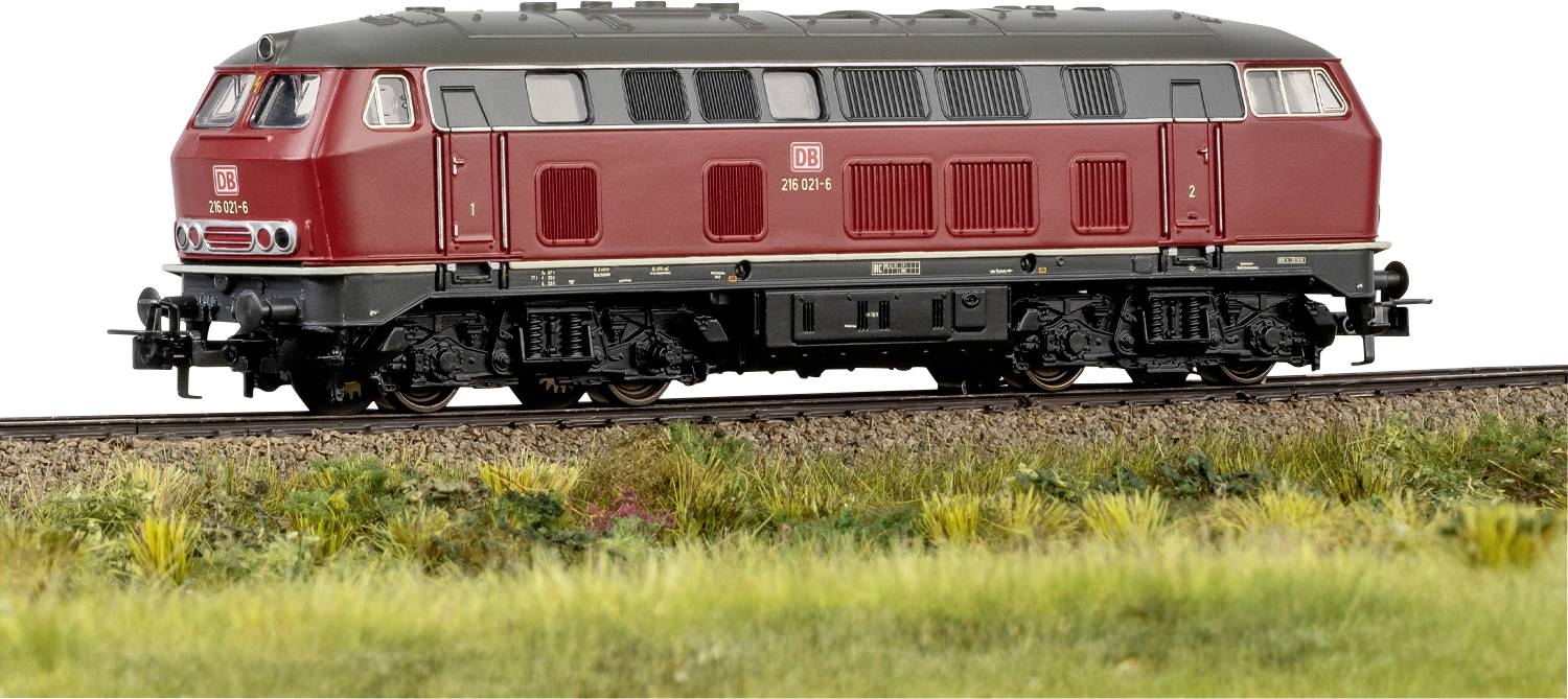 Une locomotive de train rouge étiquetée « DB 218 261-6 » sur une voie ferrée avec de l'herbe et des fleurs sauvages au premier plan, mettant en scène une scène ferroviaire rurale.