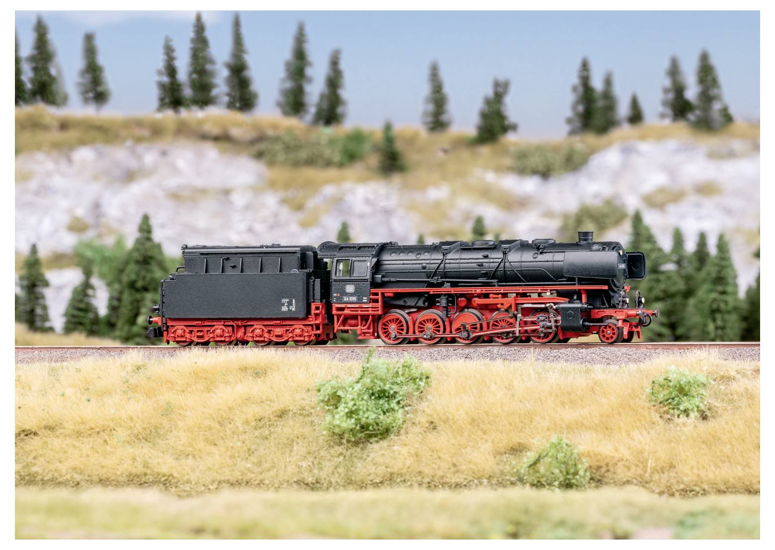 Une miniature de locomotive à vapeur avec des détails noirs et rouges est exposée sur un fond de décor représentant des arbres et une colline rocheuse.