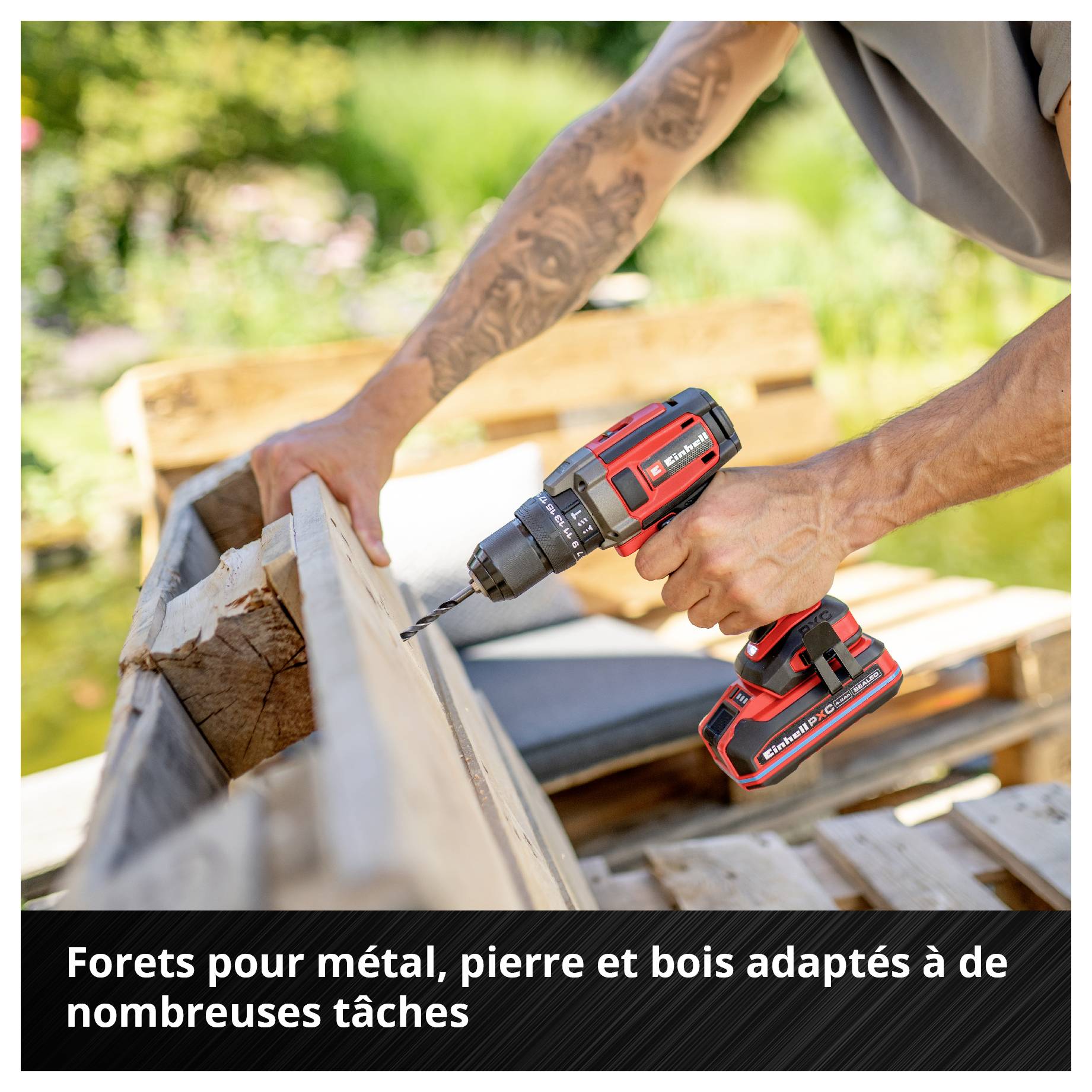 Un perceuse-visseuse sans fil rouge est utilisée par une personne pour percer du bois. En arrière-plan, des meubles de jardin flous sont visibles.
