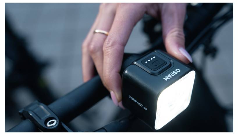 Une personne ajuste une lampe de vélo compacte montée sur le guidon, illuminant la zone devant elle. L'appareil affiche un indicateur de niveau de puissance.
