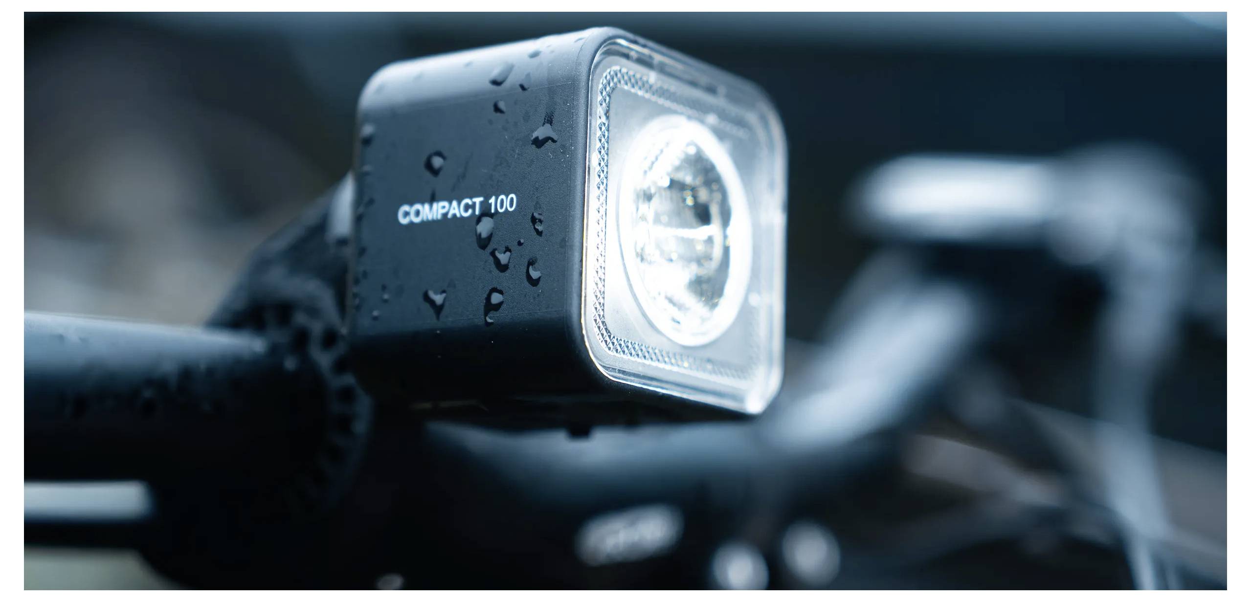Gros plan d'un phare de bicyclette étiqueté « COMPACT 100 » sur un guidon mouillé, suggérant une utilisation par temps de pluie ou par faible luminosité.