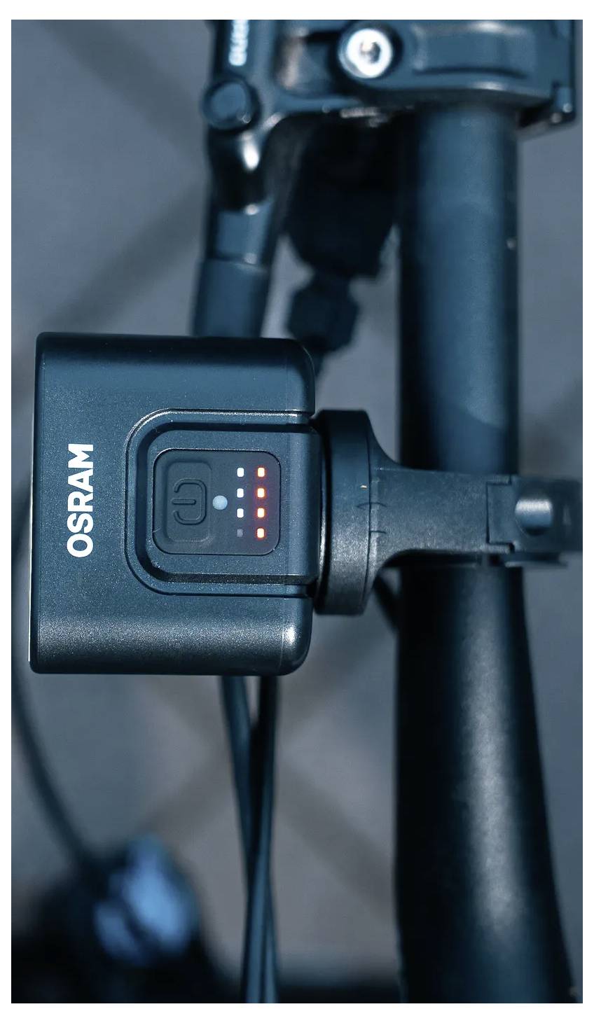 Un guidon de vélo est équipé d'un projecteur avant Osram avec quatre indicateurs LED illuminés, suggérant l'état de la batterie ou le niveau de luminosité.