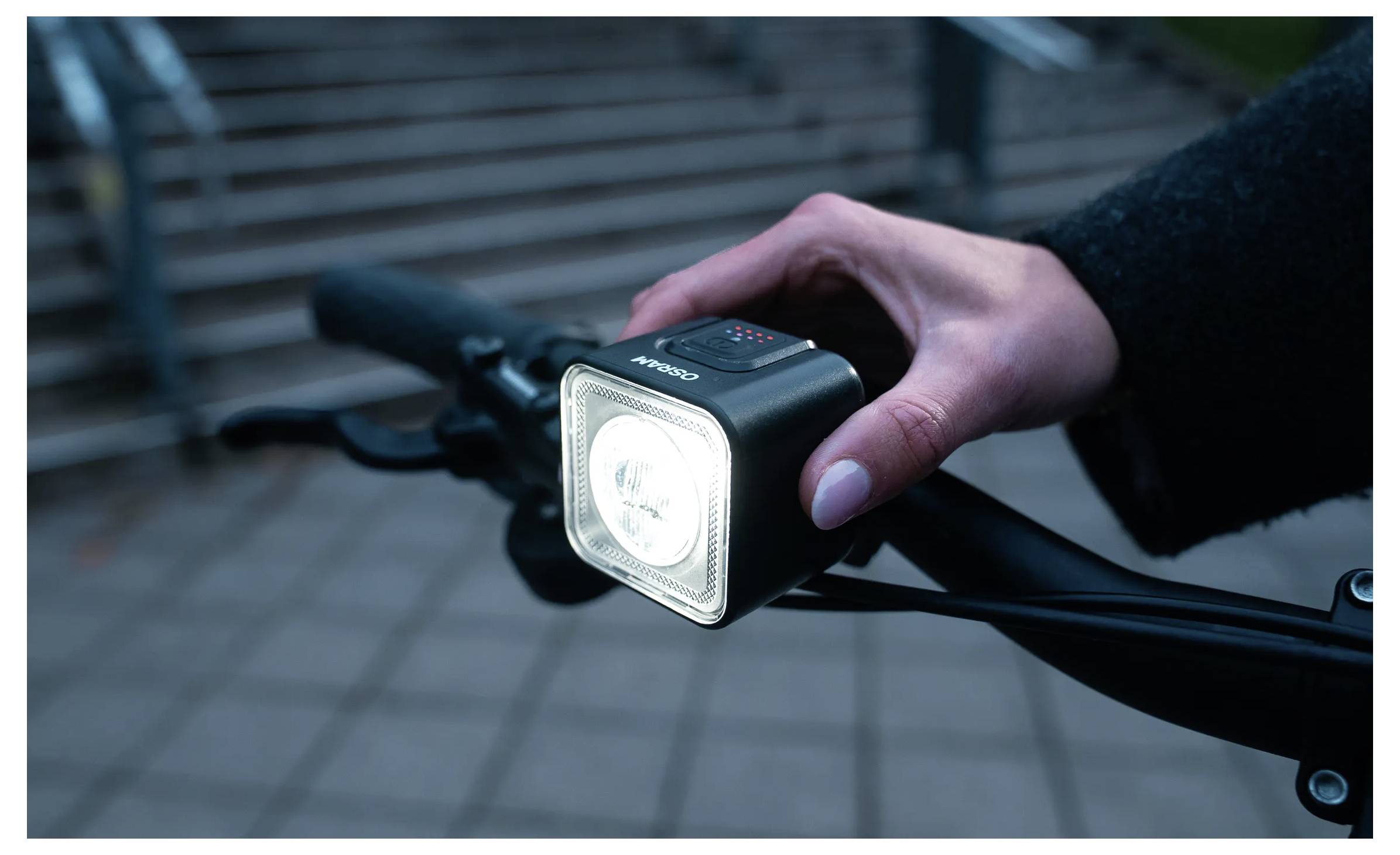 Une personne allume une lumière avant brillante sur le guidon d'un vélo, se préparant pour une balade nocturne. L'arrière-plan présente des escaliers extérieurs flous.