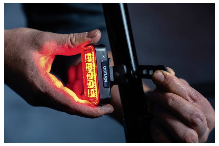 « Des mains attachant un feu de vélo Osram rouge sur le cadre d'un vélo, indiquant un équipement de sécurité pour améliorer la visibilité dans les conditions de faible luminosité. »