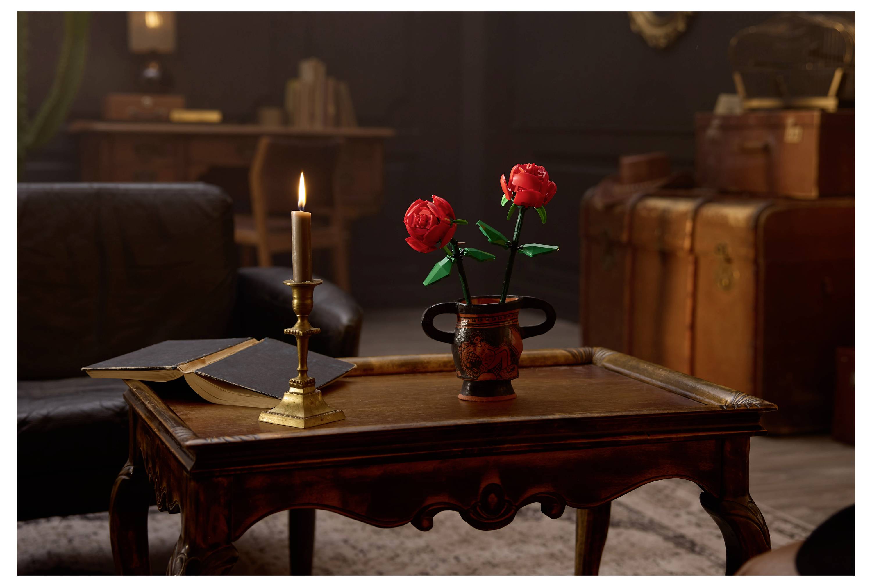 Une pièce faiblement éclairée avec une table en bois, présentant une bougie allumée, un livre ouvert et un vase de roses rouges. Des bagages vintage en arrière-plan.