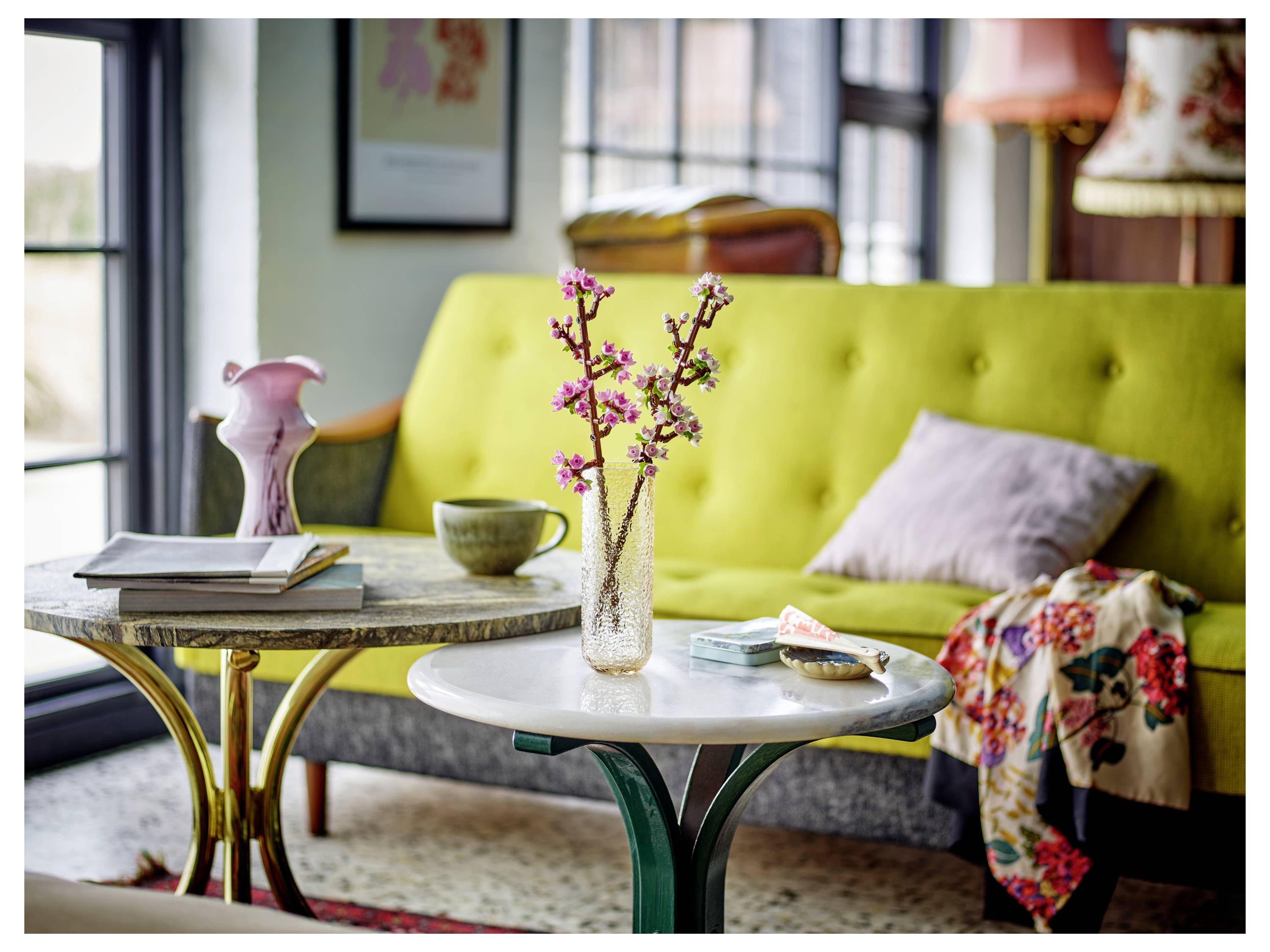 Un vase avec des fleurs de cerisier sur une petite table, disposé dans un salon avec un canapé jaune, un coussin rose et des objets décoratifs.