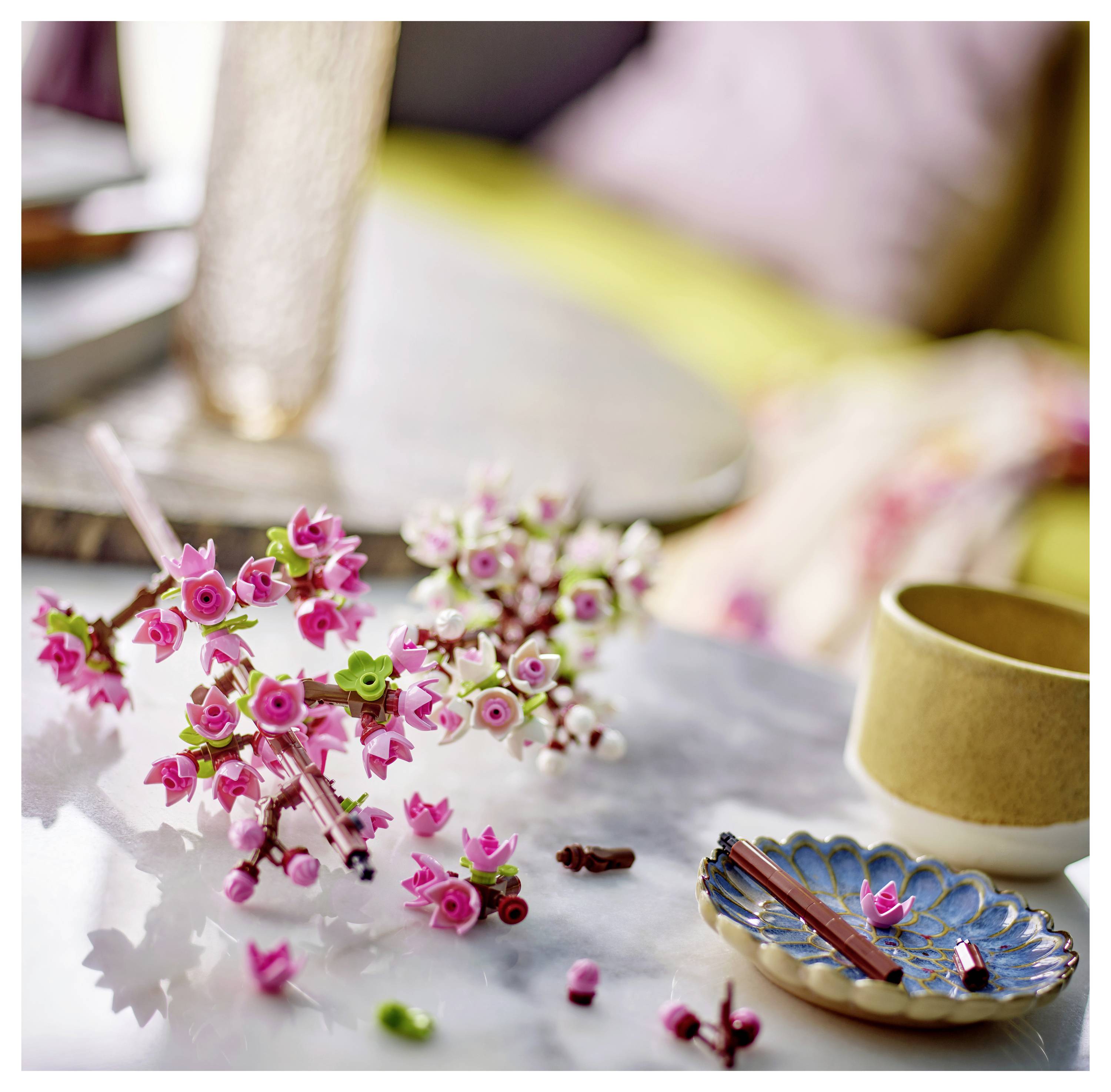 Un gros plan de petites décorations de fleurs artificielles roses et vertes sur une surface en marbre, à côté d'une tasse beige et d'une assiette décorée.