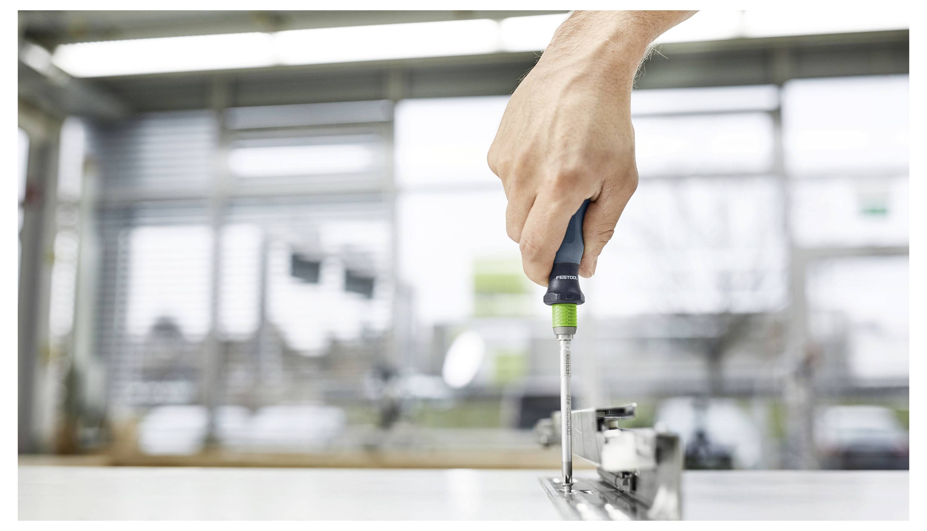 Une main utilise un tournevis pour assembler ou ajuster un morceau de quincaillerie métallique dans un environnement industriel lumineux.