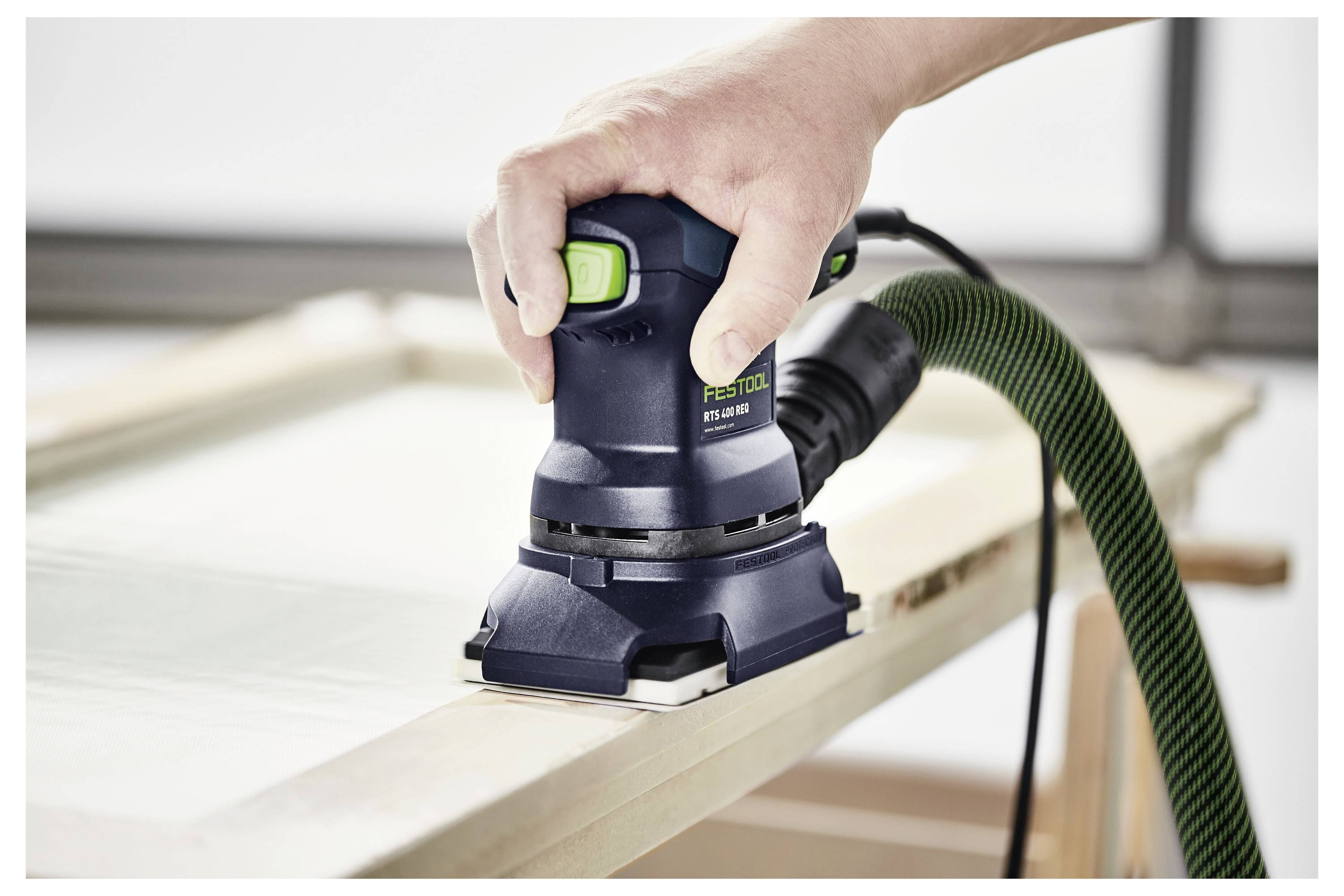 Une main actionne une ponceuse électrique sur une surface en bois, en lissant les arêtes. La ponceuse est connectée à un tuyau vert pour l'extraction de la poussière.