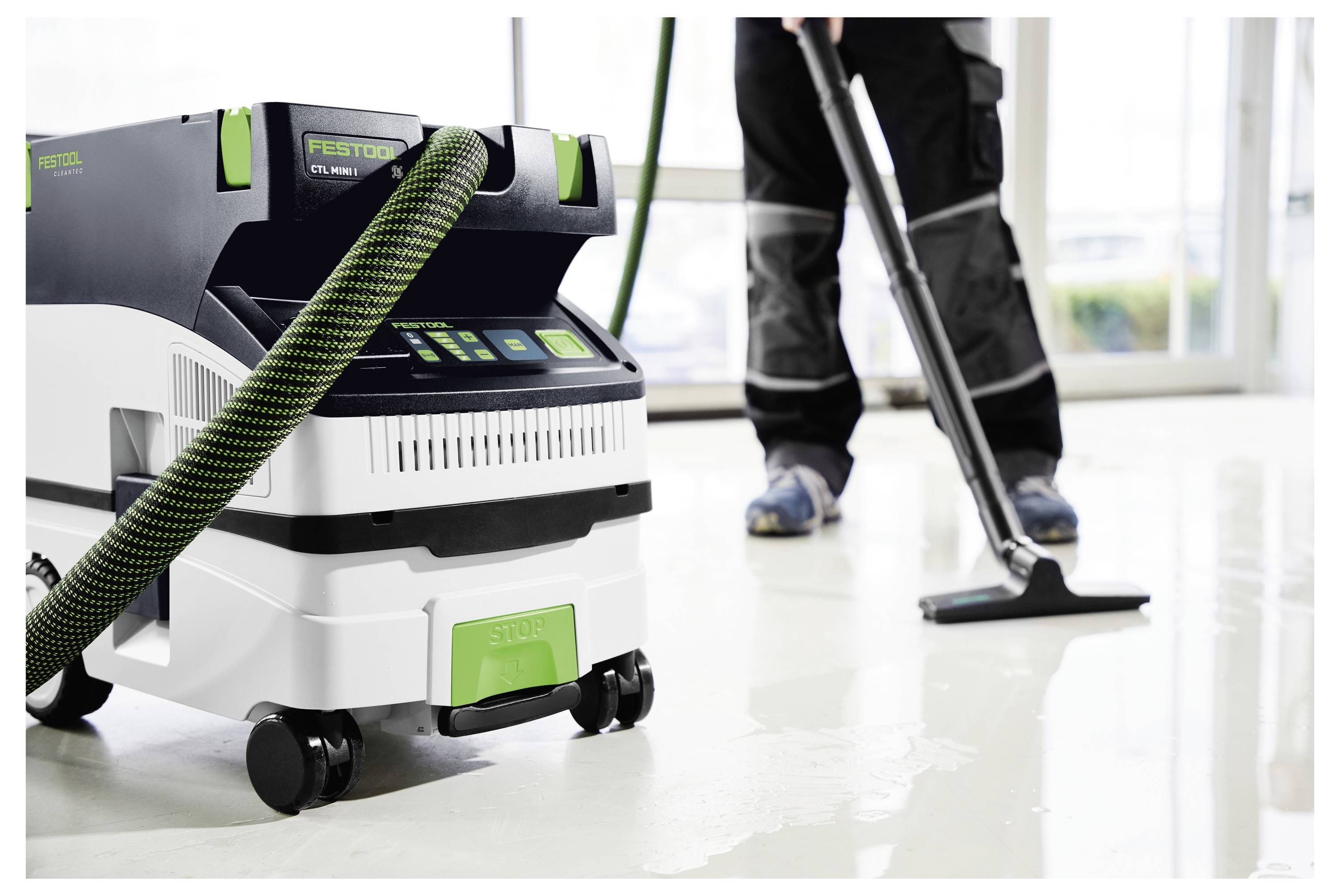 A person in work pants uses a floor cleaning machine with a vacuum attachment to clean a shiny, light-colored floor indoors.