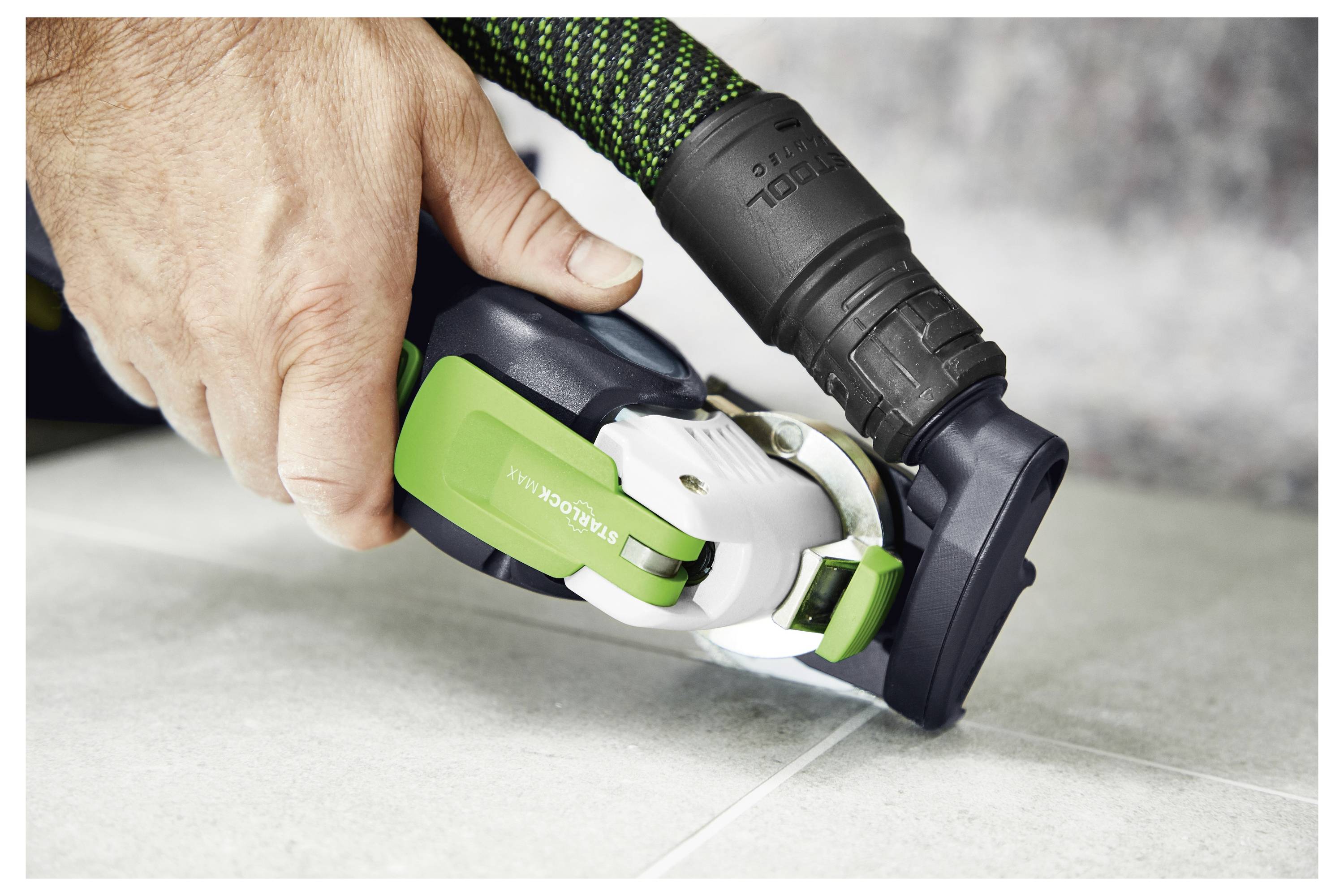 Une personne utilise un outil électrique avec un protecteur vert pour couper des carreaux, avec un système d'aspiration collectant la poussière près de la zone de coupe.