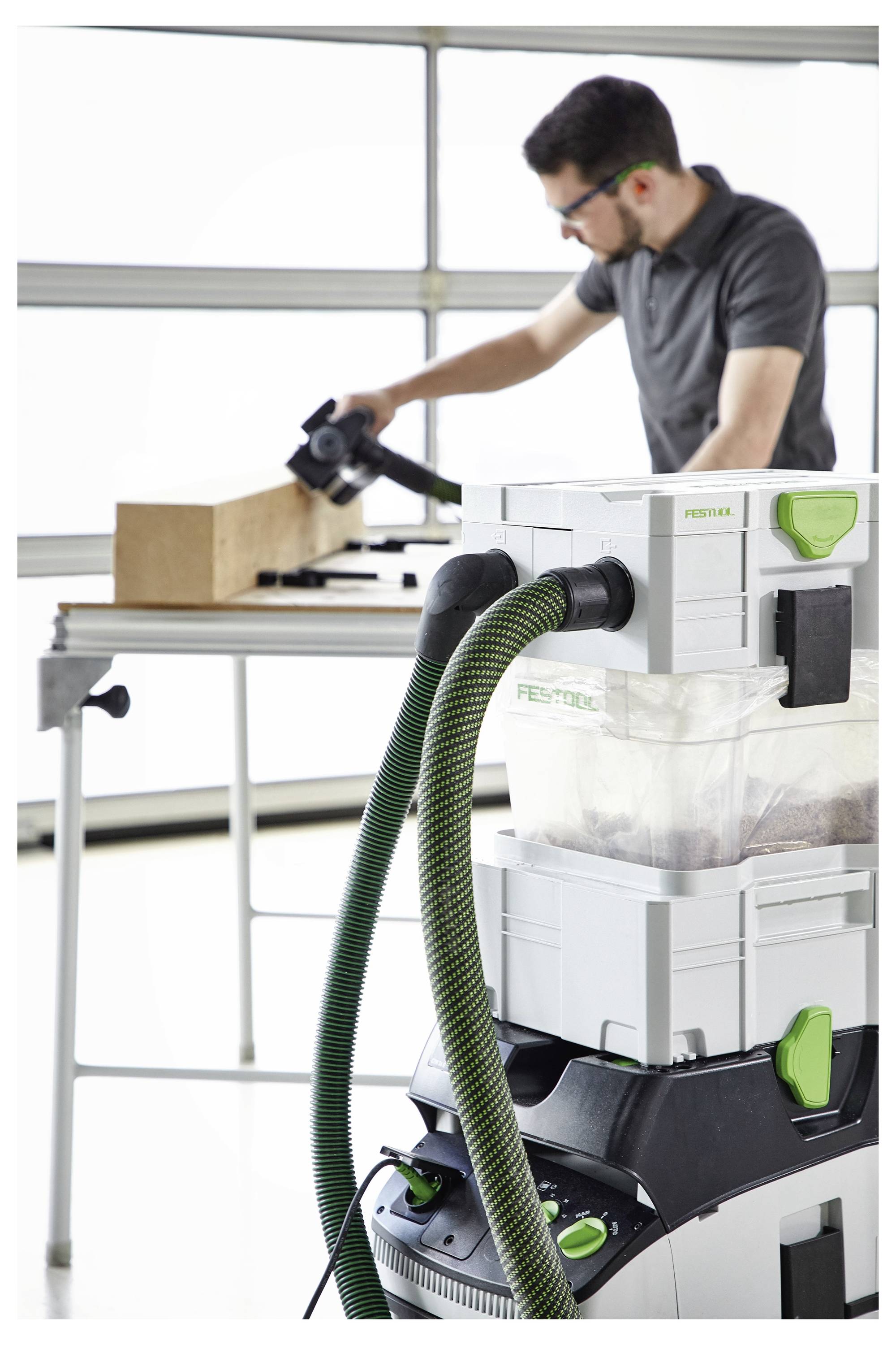 'Une personne poncant un bloc de bois sur un établi, avec un aspirateur extracteur de poussière connecté au premier plan, à l'intérieur d'un atelier.'
