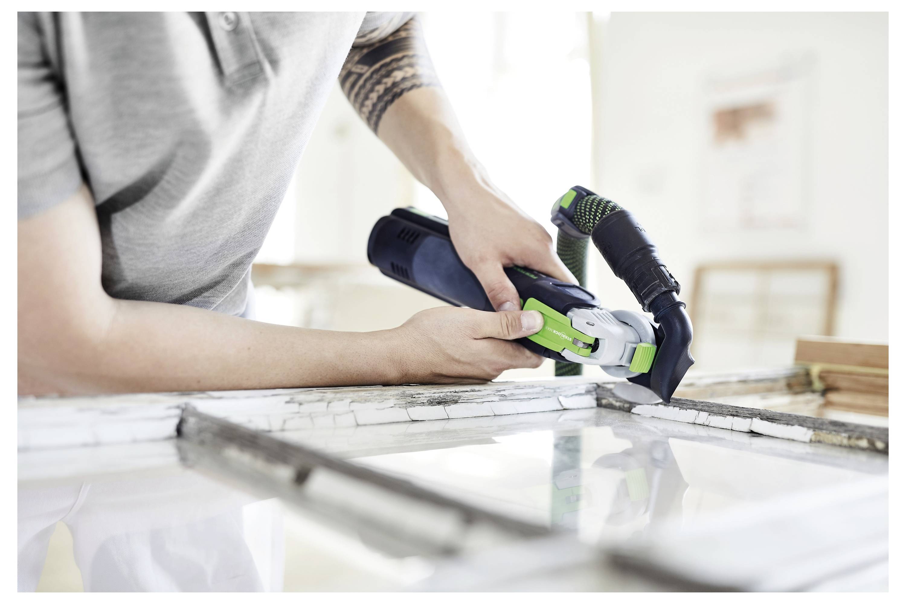 Une personne vêtue d'une chemise grise utilise un outil électrique noir et vert pour découper un cadre de fenêtre, travaillant sur un projet de rénovation domiciliaire.