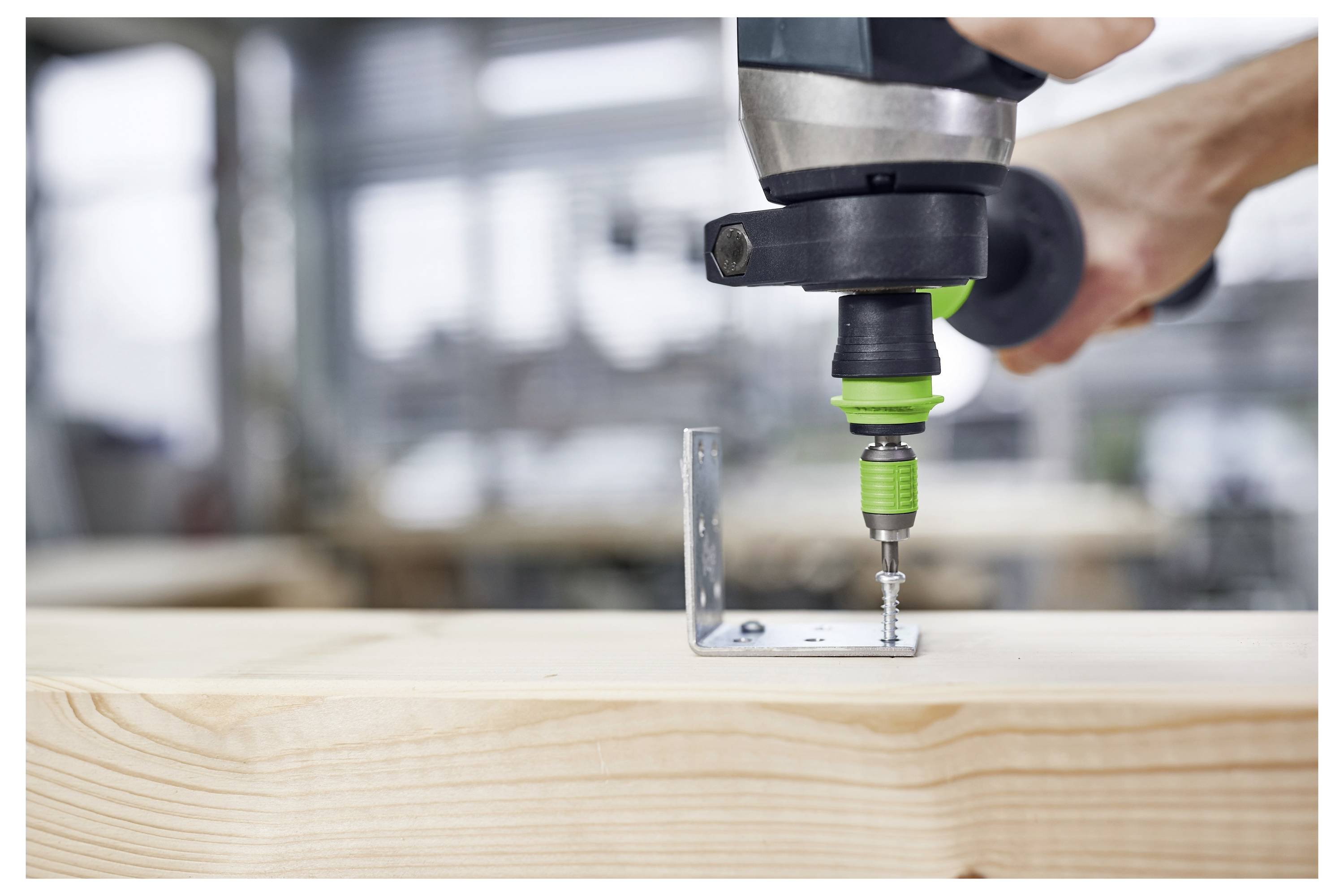 Une personne utilise une perceuse à poignée verte pour visser un support métallique sur une planche de bois dans un atelier.