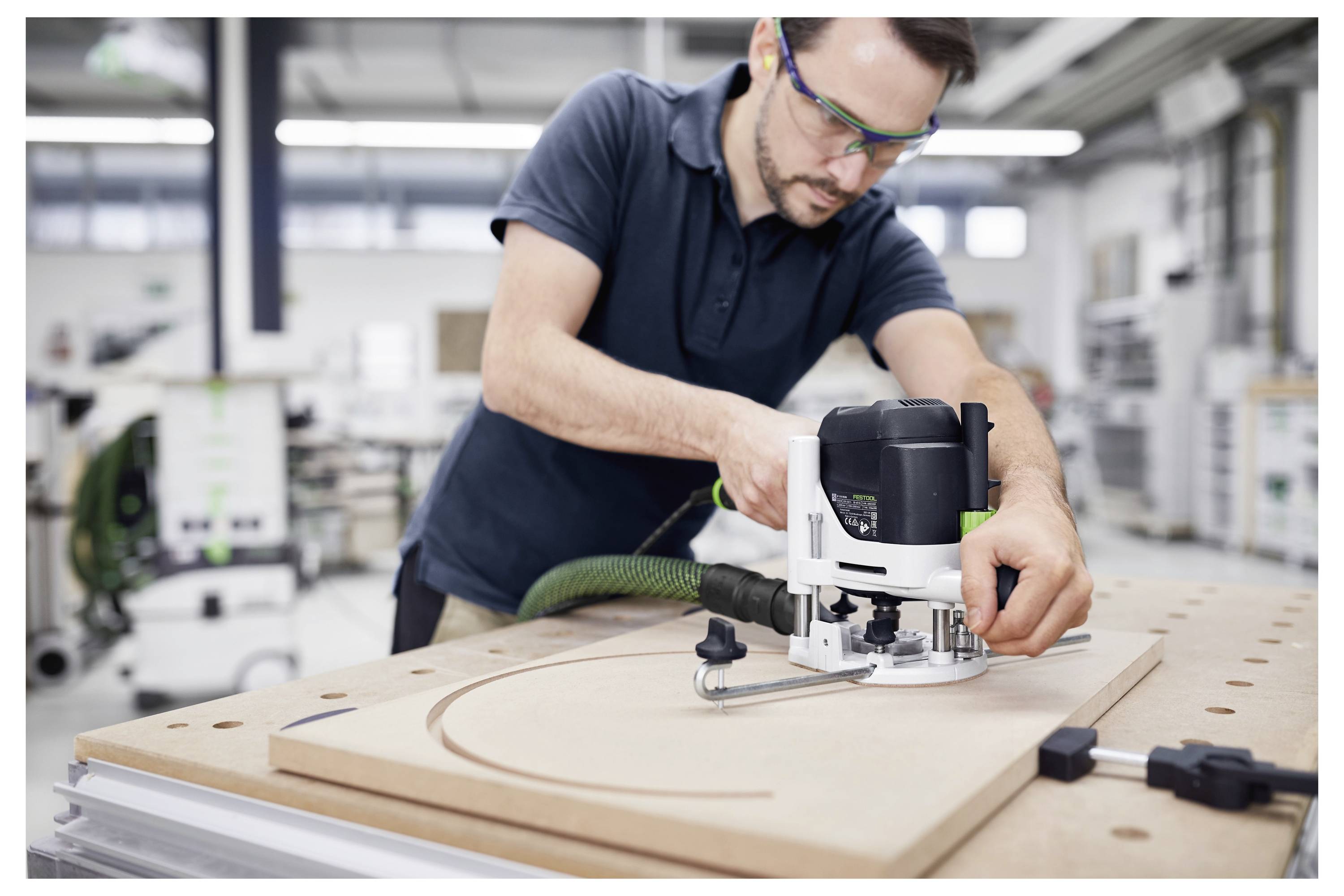 A person uses a router tool to carve a curved line into a wooden board in a workshop, wearing safety glasses and casual attire.