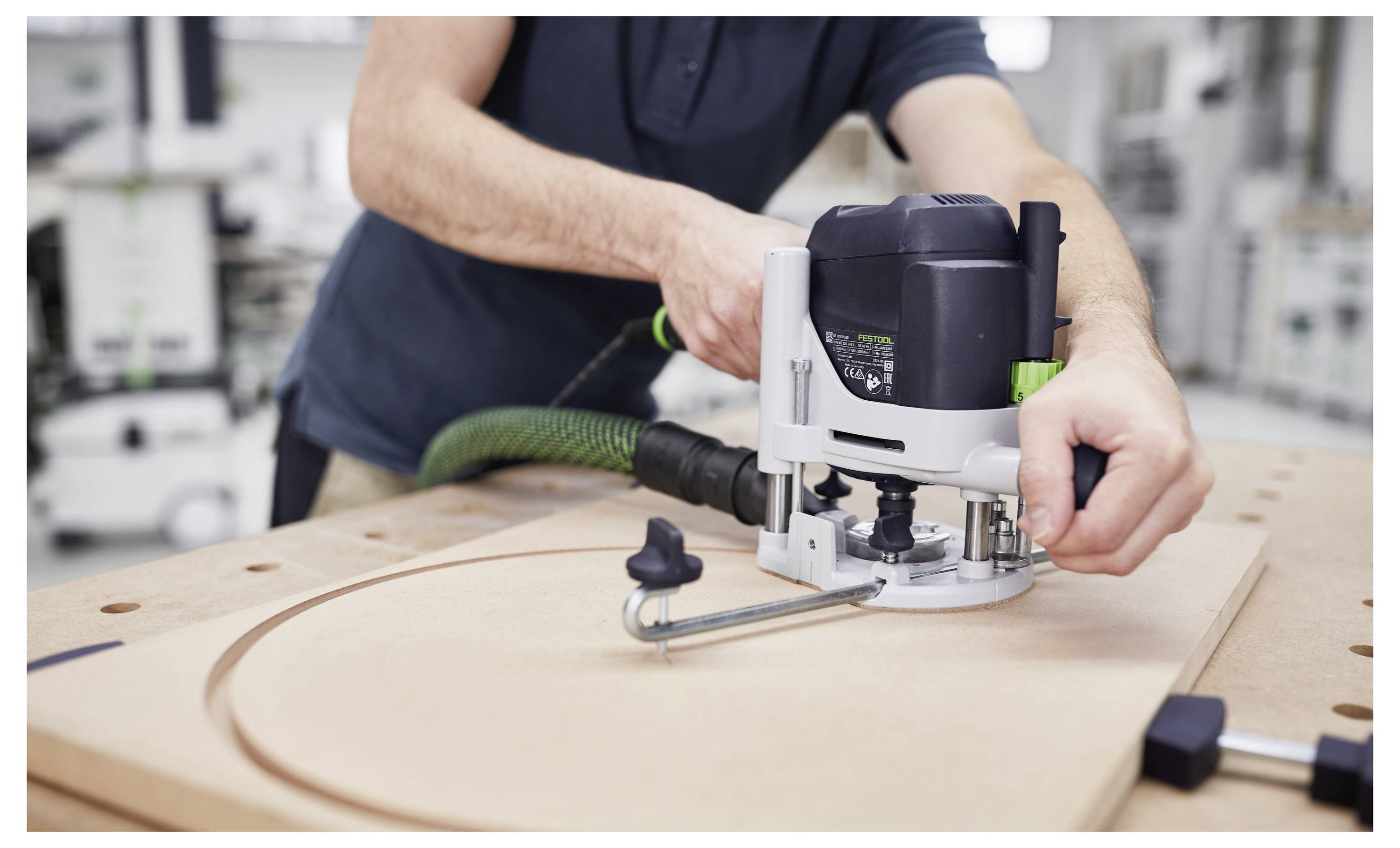 Un routeur Festool monté sur une table de travail coupant une pièce de bois dans un atelier, avec un tuyau d'aspiration attaché pour l'extraction de la poussière.
