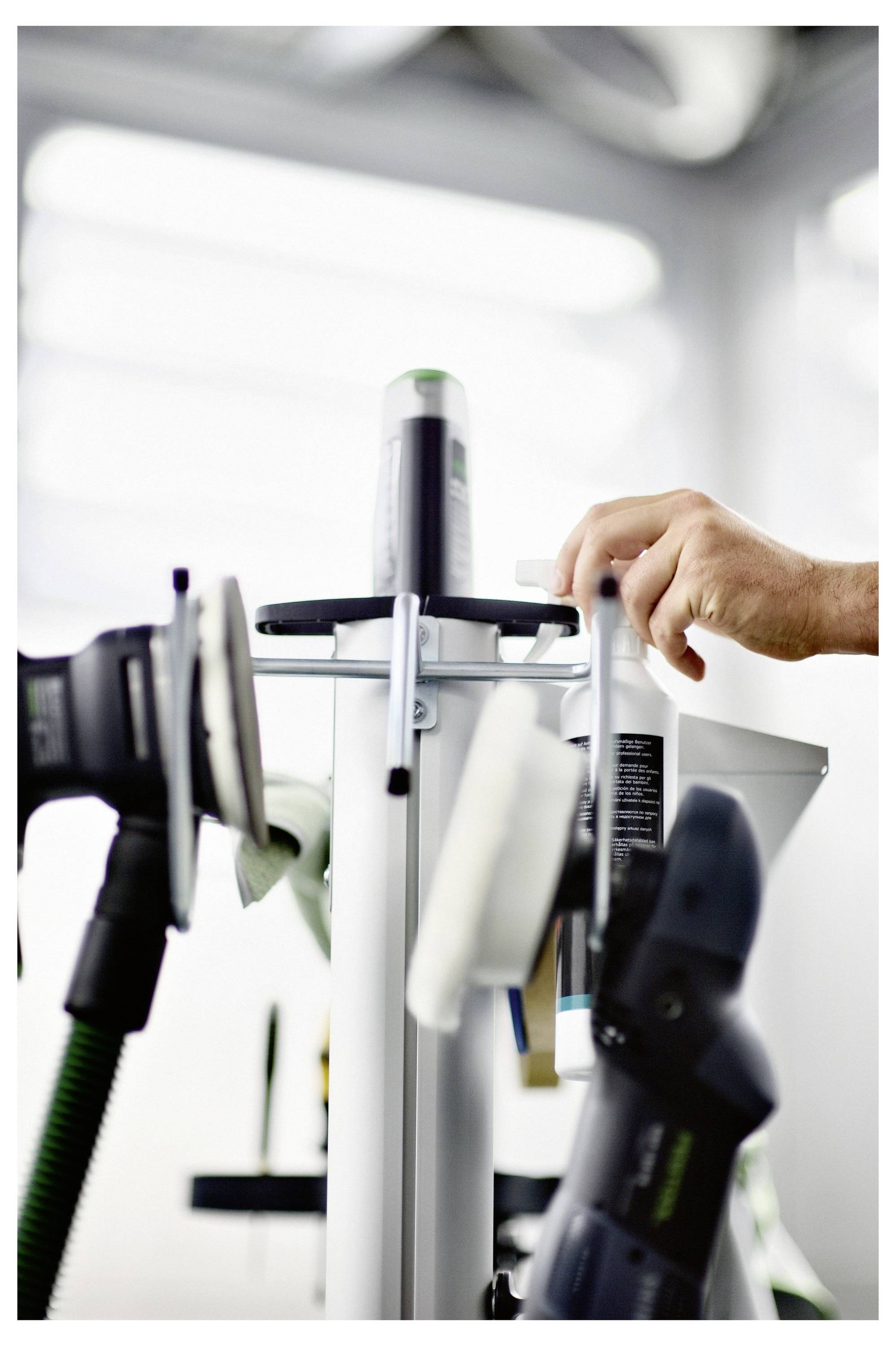 La main d'une personne ajuste des outils sur un support dans un atelier, ce qui suggère la préparation d'une tâche ou d'un projet. Les outils incluent des ponceuses et un rouleau.