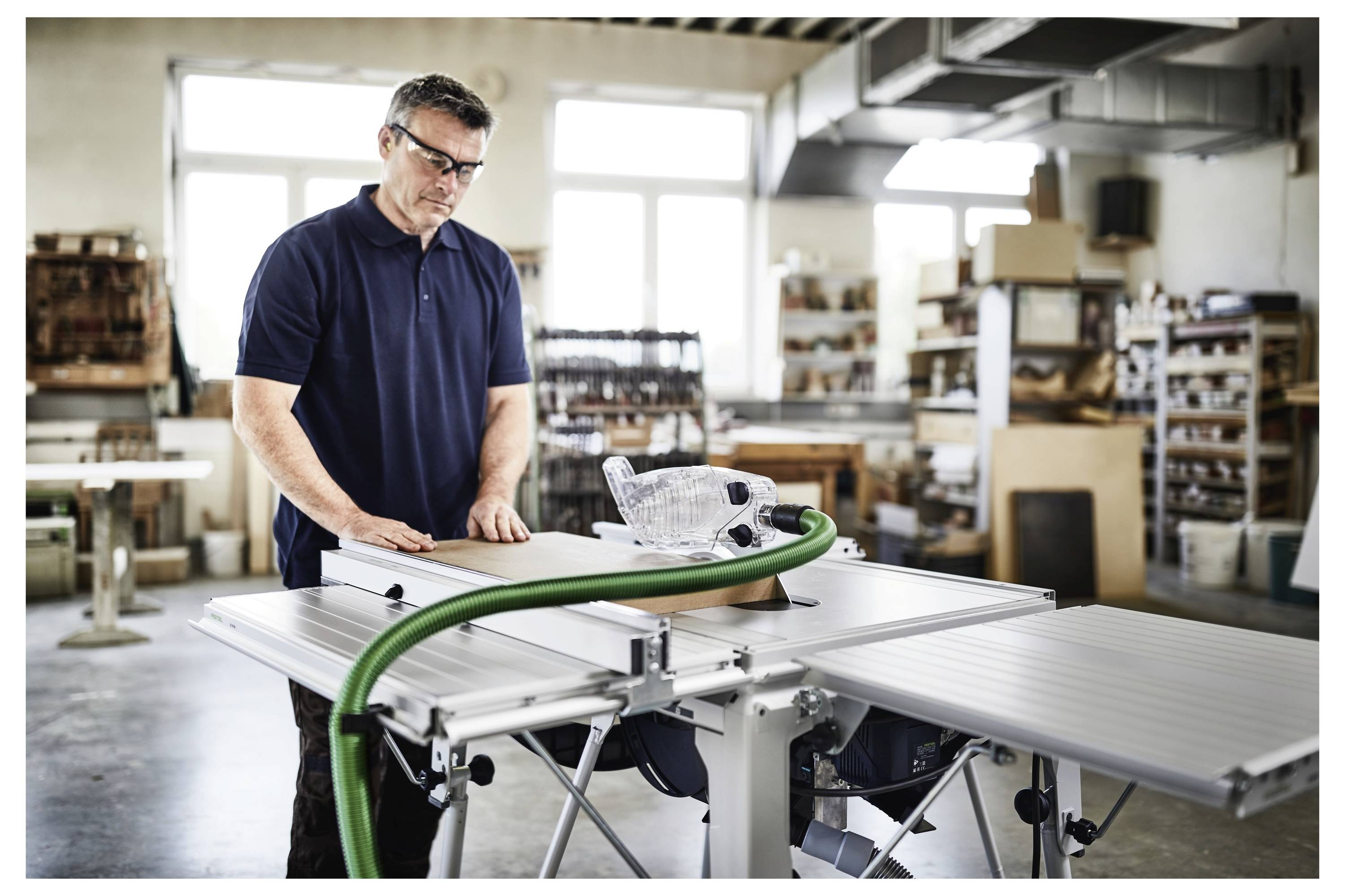 Une personne portant des lunettes de sécurité utilise une scie circulaire de table dans un atelier, entourée d'étagères et d'outils de menuiserie.