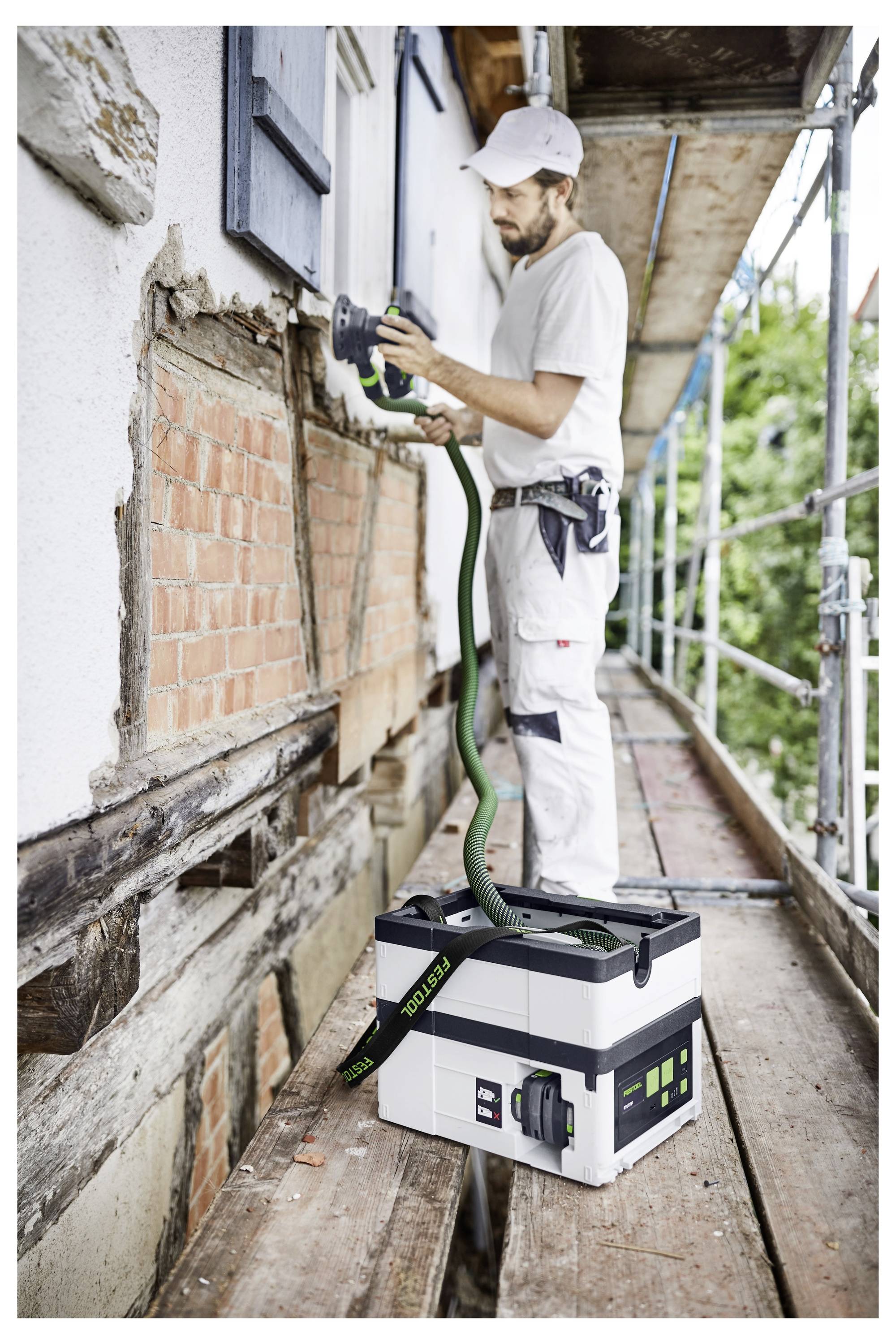 Un ouvrier de la construction utilise un outil électrique sur le mur extérieur d'un bâtiment, se tenant sur un échafaudage. Une boîte à outils se trouve sur la plateforme de l'échafaudage.