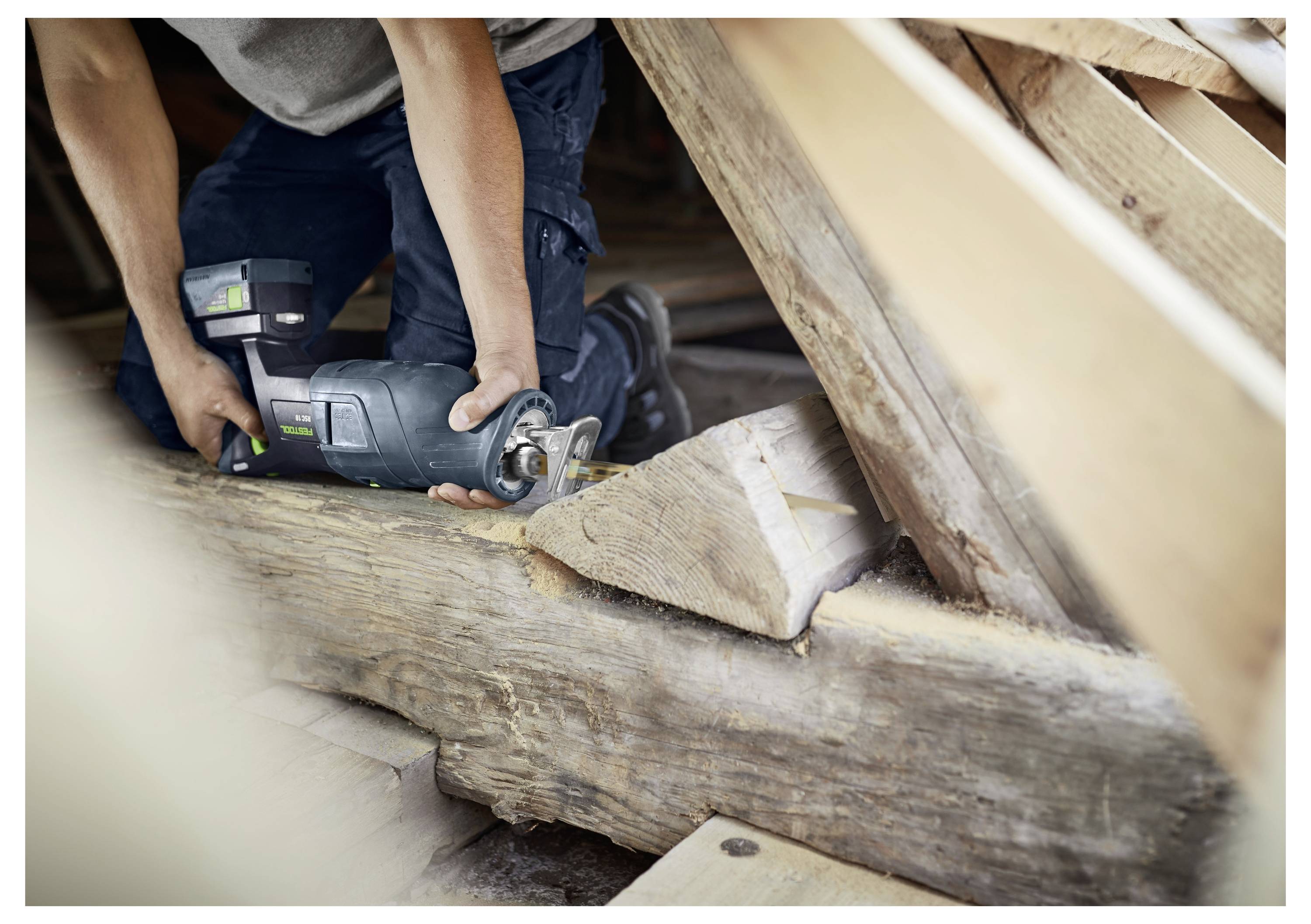 A person uses a power saw to cut a wooden beam in an attic space with visible rafters, suggesting construction or renovation work.