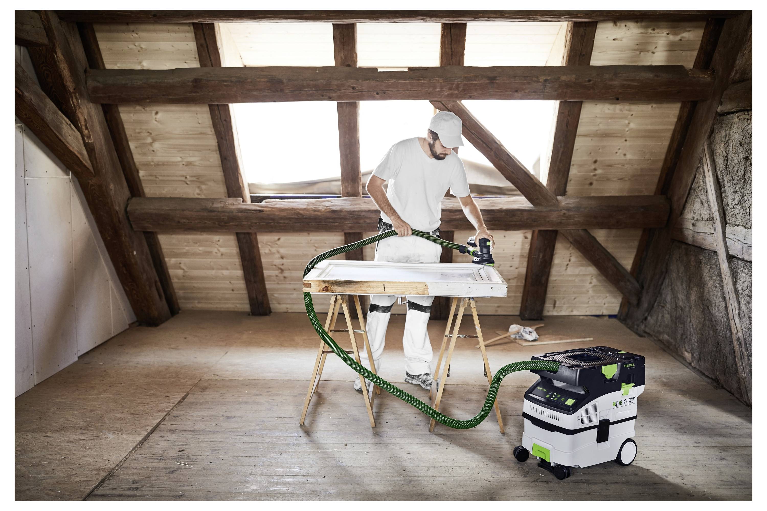 Une personne dans un grenier utilise un outil électrique sur du bois supporté par des tréteaux, connecté à un extracteur de poussière. Des poutres en bois et des murs sont visibles.