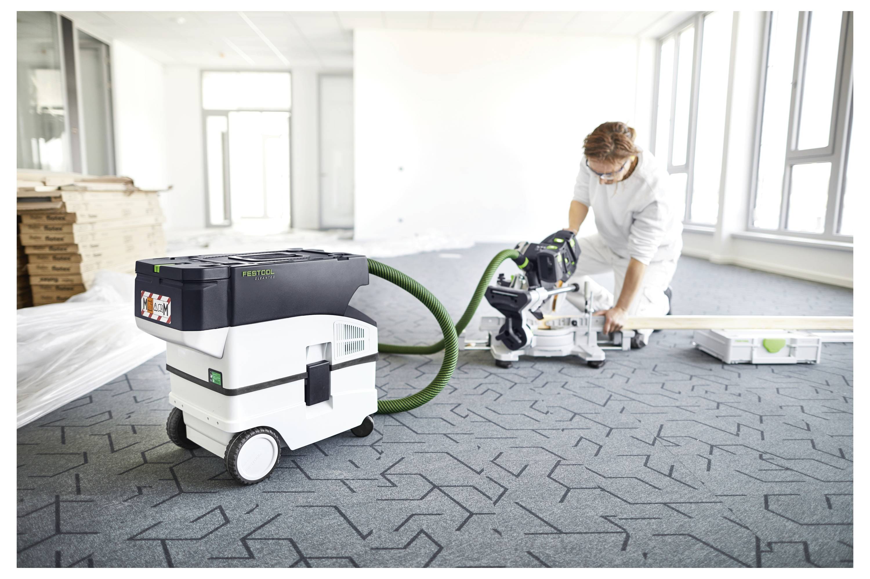 Une personne utilise un outil électrique connecté à un aspirateur dans une grande pièce lumineuse avec des équipements non emballés et des matériaux emballés à proximité.