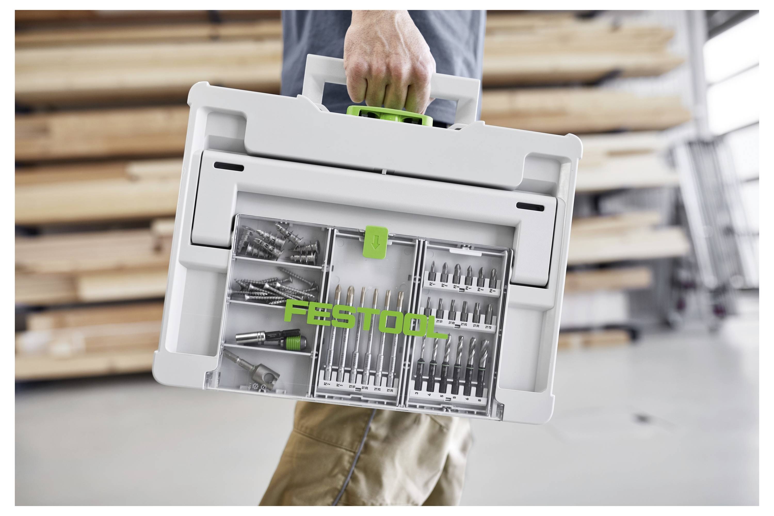 Person carrying a gray Festool tool case with a green latch, containing organized drill bits and accessories in a workshop setting.