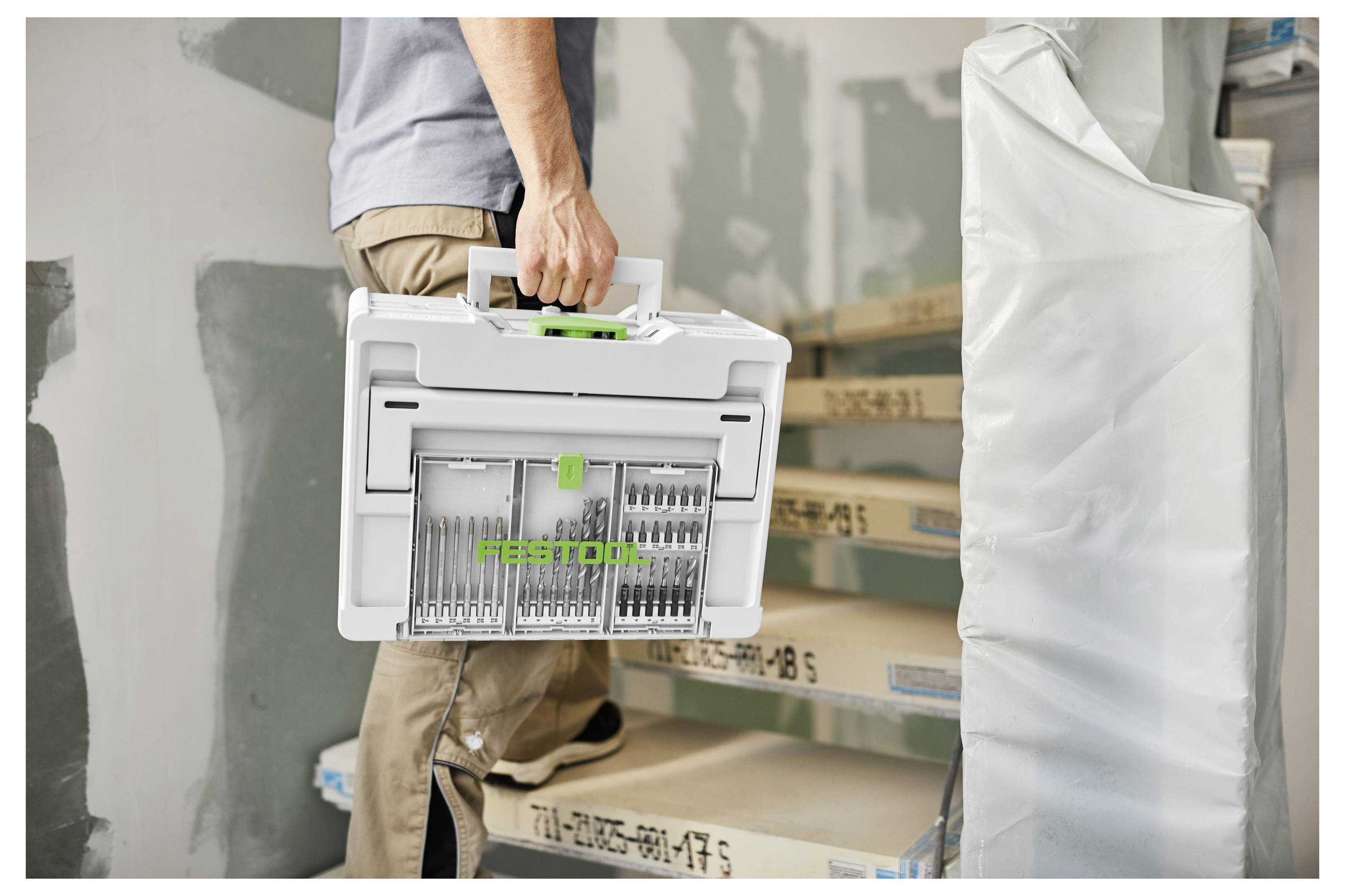 Une personne portant un grand conteneur de rangement blanc avec des compartiments en montant un escalier partiellement construit, indiquant des travaux de construction.