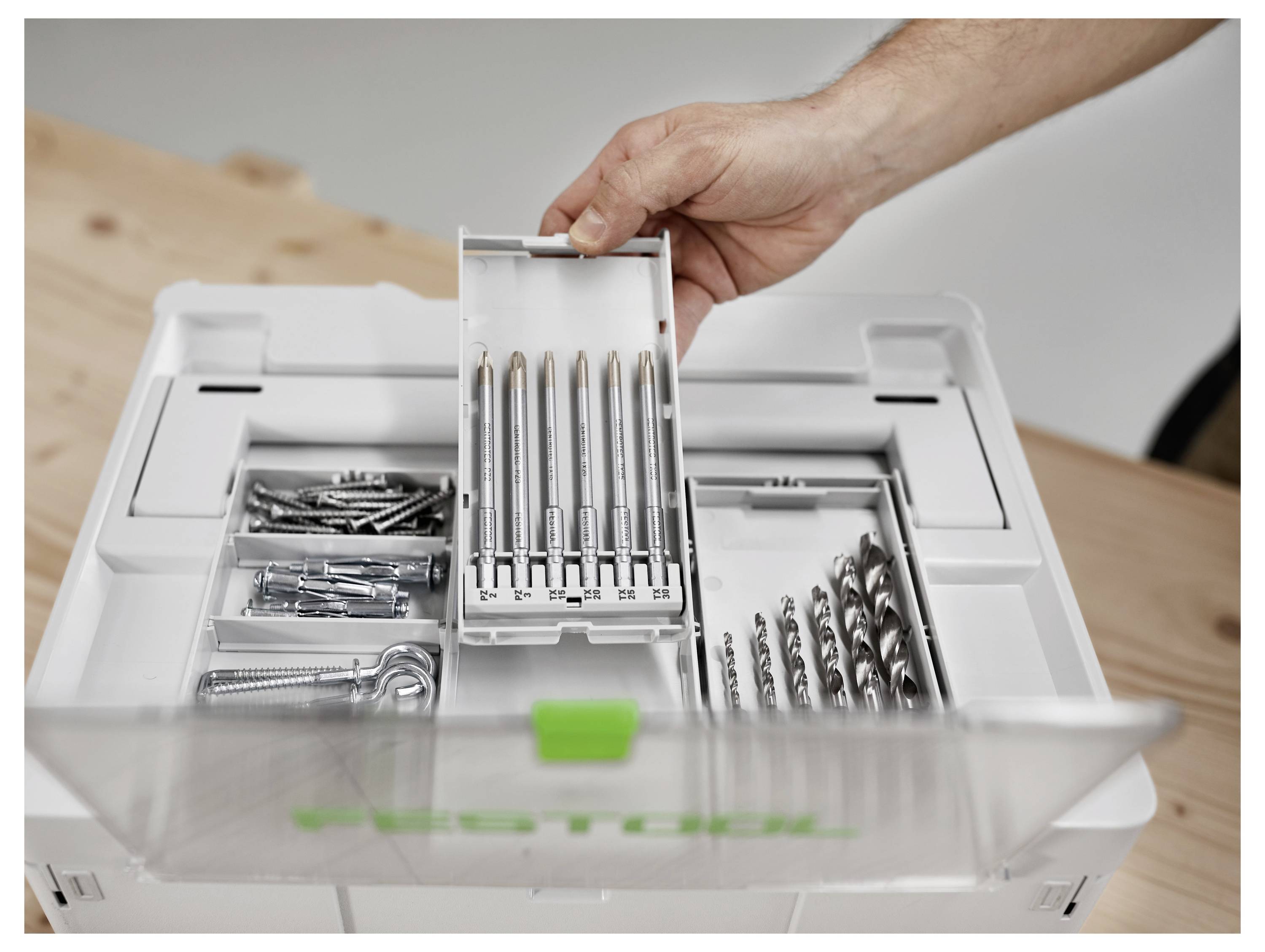 Une main sélectionnant un ensemble de mèches de perceuse et de embouts de tournevis dans une boîte à outils organisée, mettant en évidence diverses fixations et accessoires dans une boîte blanche.