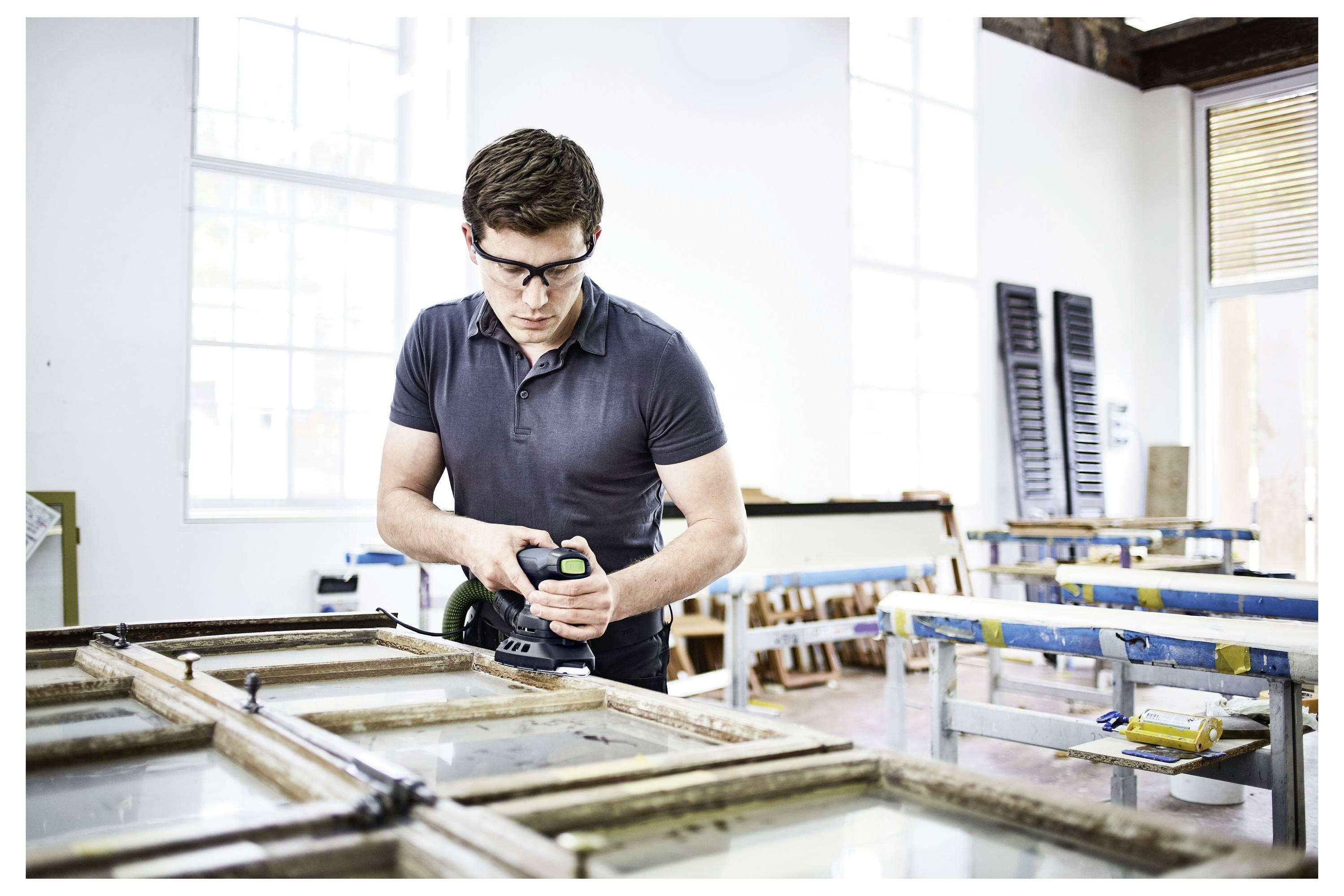 Une personne dans un atelier rénovant des cadres de fenêtres avec des outils, portant des lunettes de sécurité et un polo. Espace de travail lumineux avec de grandes fenêtres.