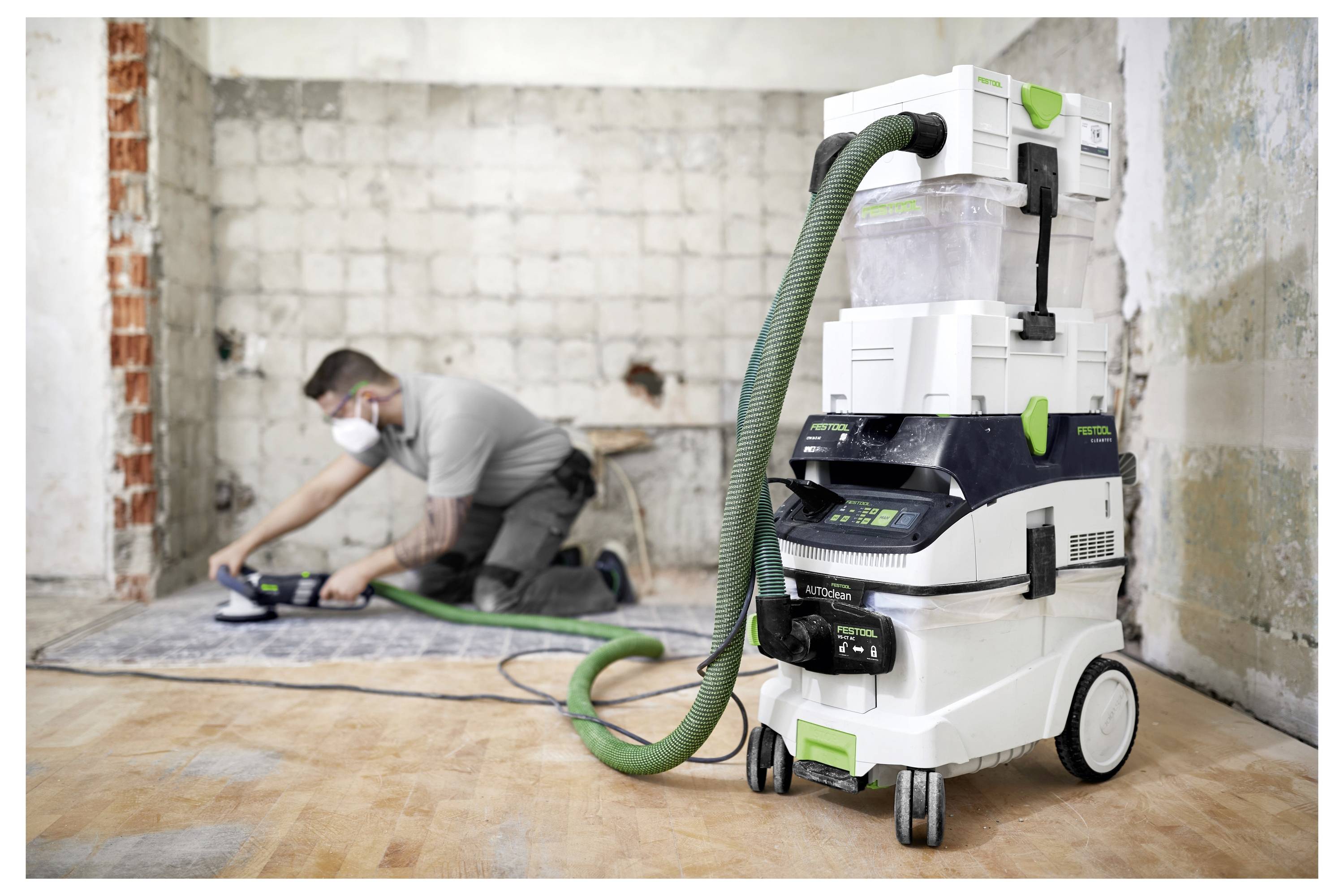 Un travailleur portant un masque utilise un outil électrique près d'un mur dans une pièce en rénovation. Un aspirateur avec un tuyau vert se trouve au premier plan.