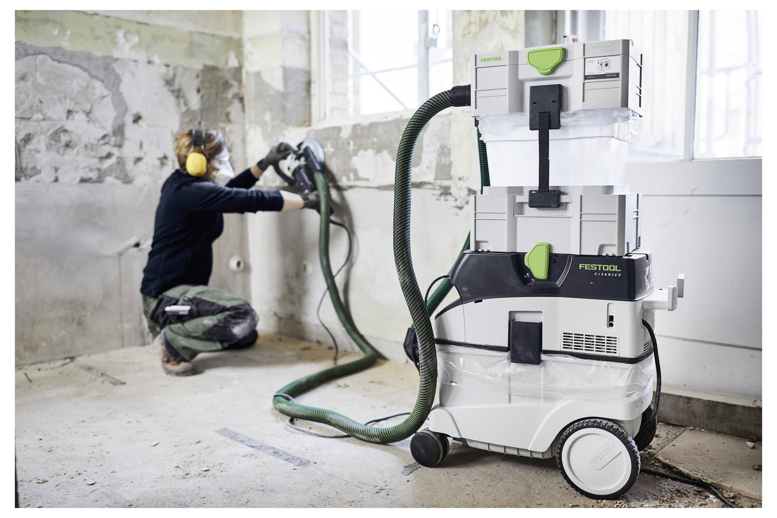 Une personne vêtue d'équipement de protection fait fonctionner une ponceuse murale à côté d'un extracteur de poussière Festool dans une pièce partiellement rénovée.