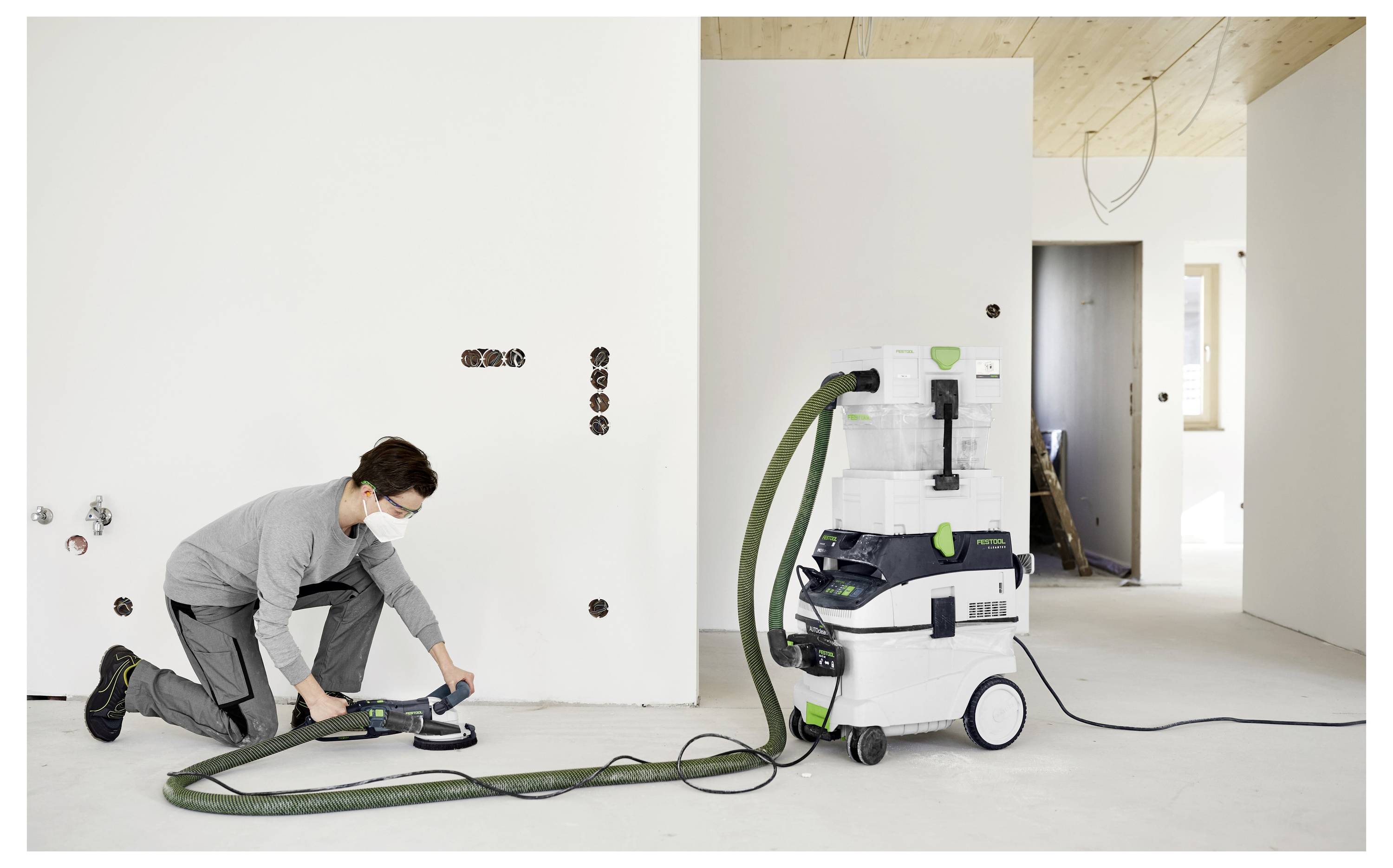 Une personne utilise un outil électrique sur un mur dans une pièce partiellement terminée. Un système d'aspiration est connecté à l'outil pour collecter la poussière.