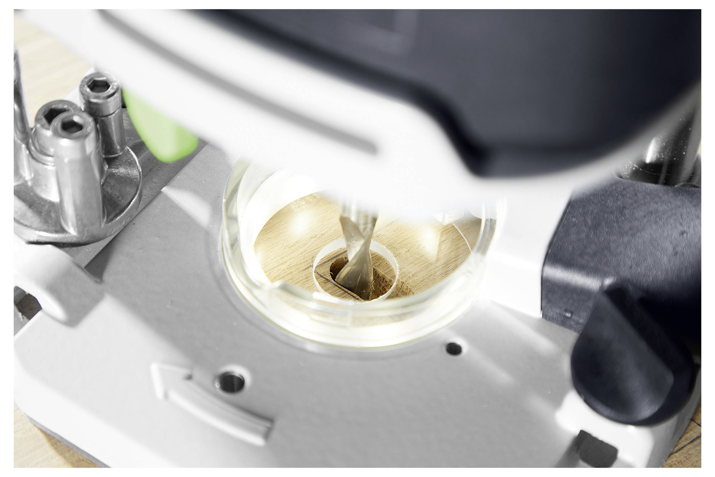 A drill bit is actively cutting through a wooden surface, mounted on a mechanical base with metal and plastic components.