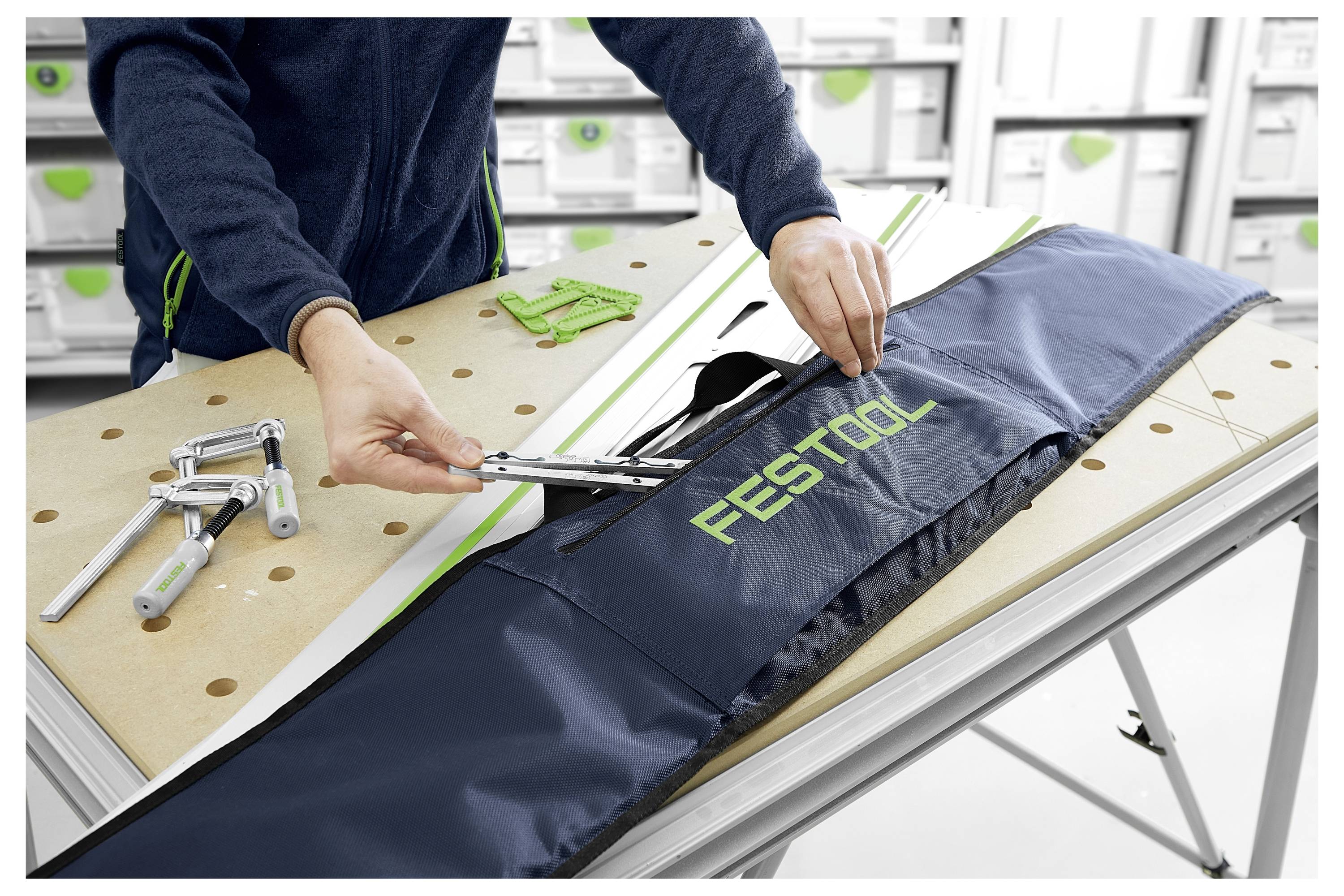 Une personne insère des outils dans un sac de marque « Festool » sur un établi, entourée d'autres outils et de boîtes de rangement dans un atelier.