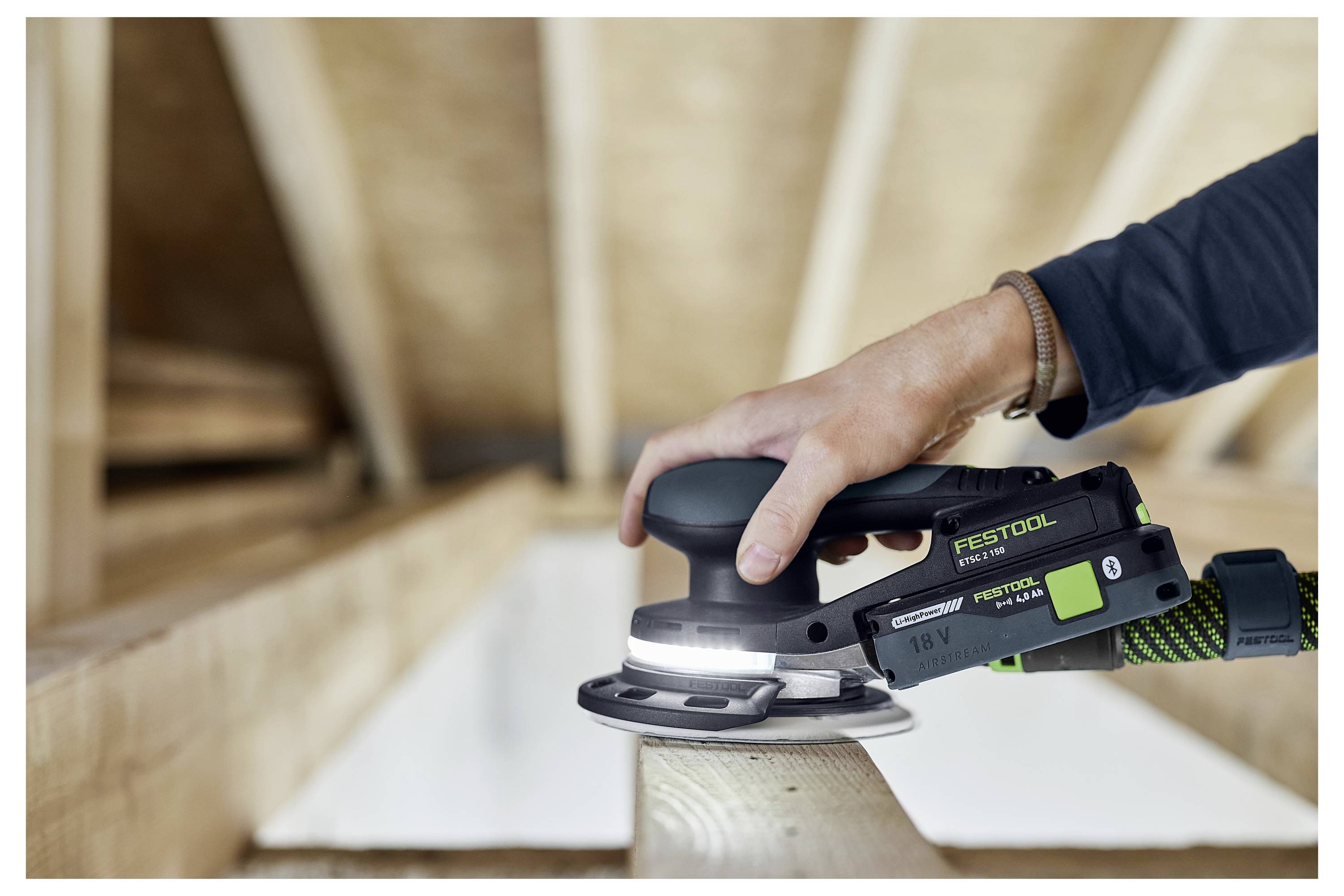 A person uses a handheld circular sander on a wooden beam, surrounded by an unfinished wooden framework.