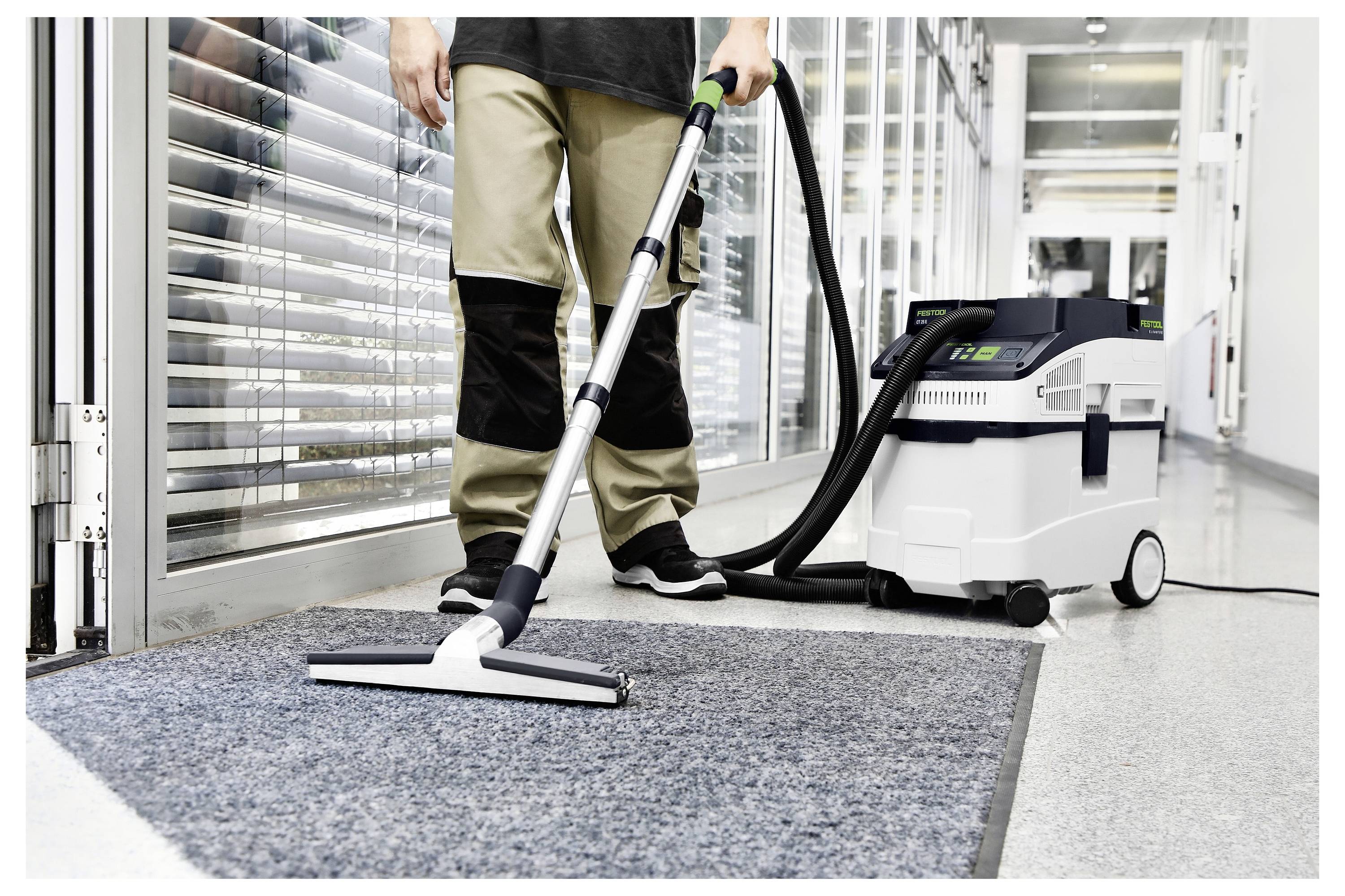 « Une personne passant l'aspirateur sur le tapis d'un couloir avec un aspirateur commercial, portant un pantalon de travail noir et kaki et des chaussures noires, près de grandes fenêtres. »