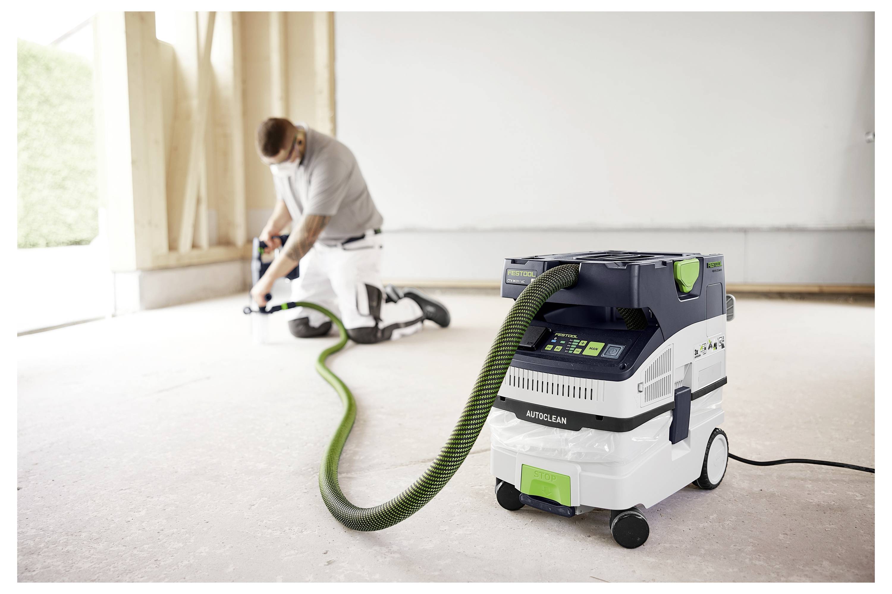 A person kneels on a construction site using a power tool connected to a dust extractor. The device is central, highlighting dust control.