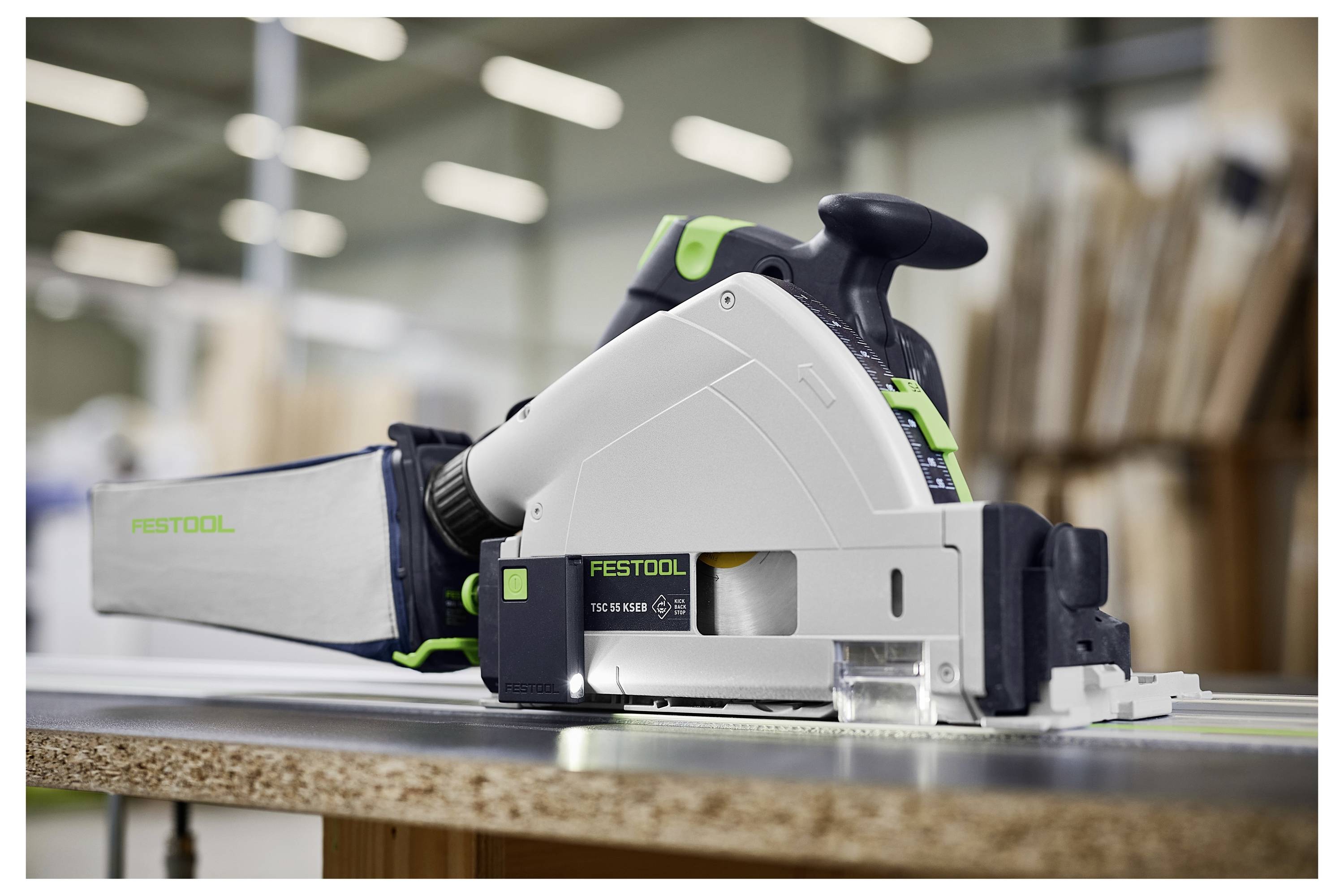 Une scie à rail Festool repose sur une planche de bois dans un atelier, prête pour une coupe précise. L'arrière-plan flou suggère un espace de travail actif.