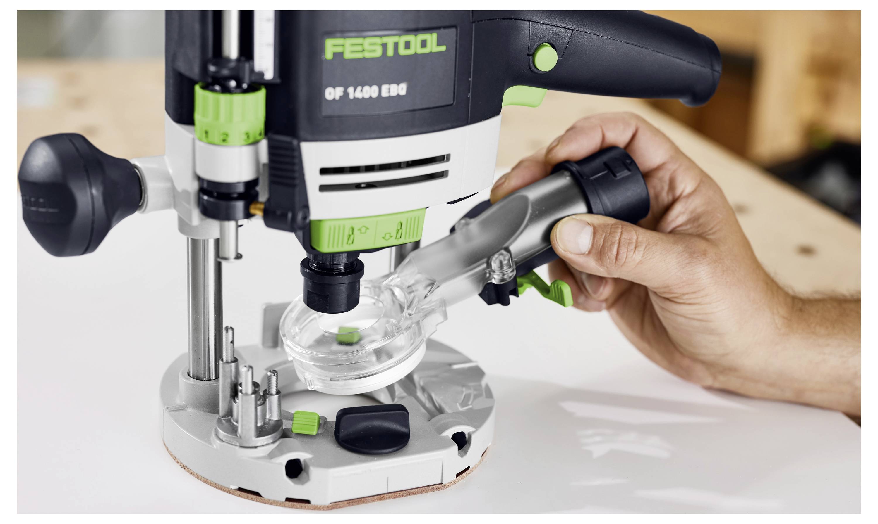 Une main ajuste un accessoire d'extraction de poussière transparent sur une toupie Festool OF 1400 EBQ placée sur une surface en bois, démontrant la mise en place de l'outil.