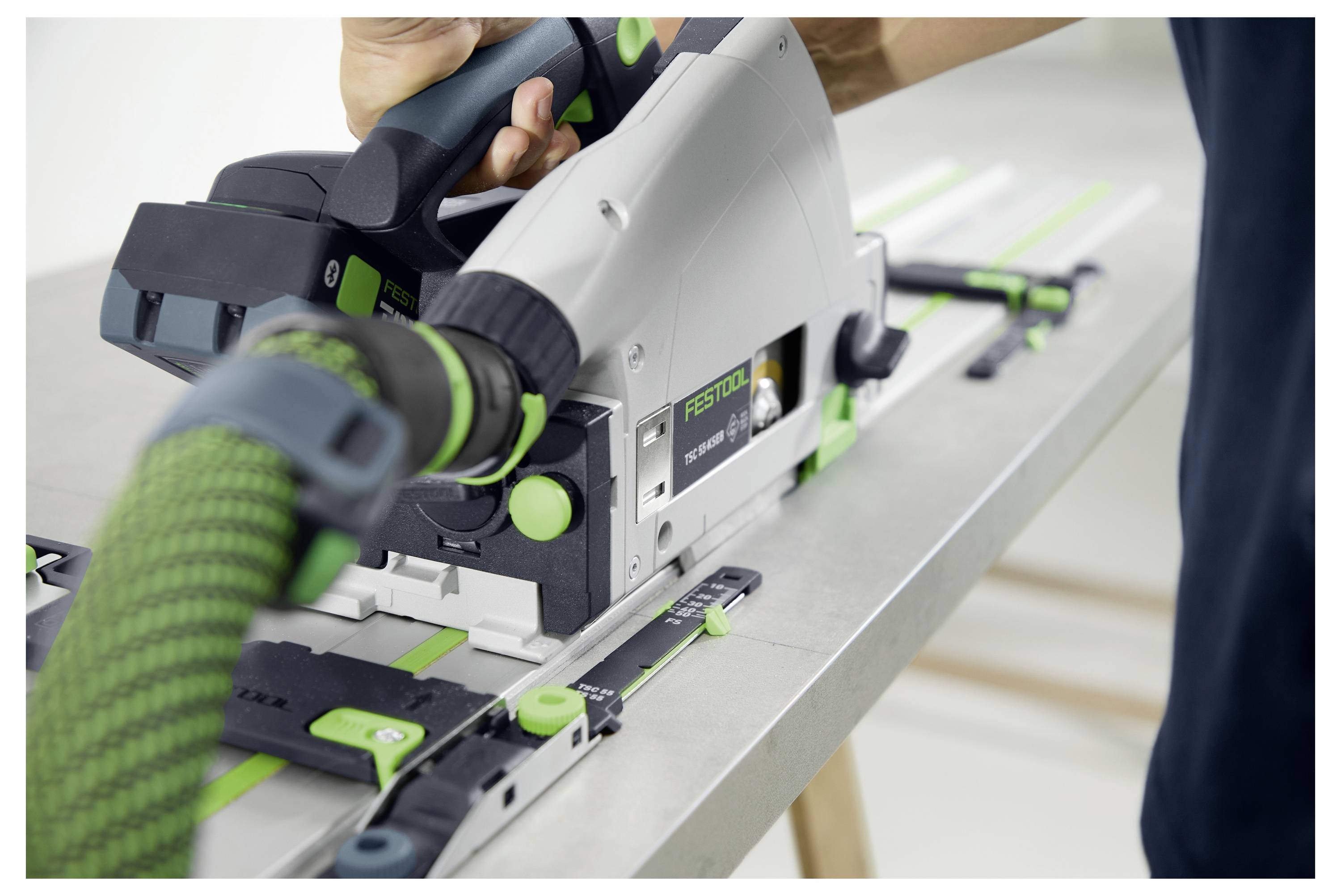 Une personne utilisant une scie circulaire à piste Festool pour couper une planche, avec un tuyau d'aspiration vert et noir connecté pour la collecte de la poussière.