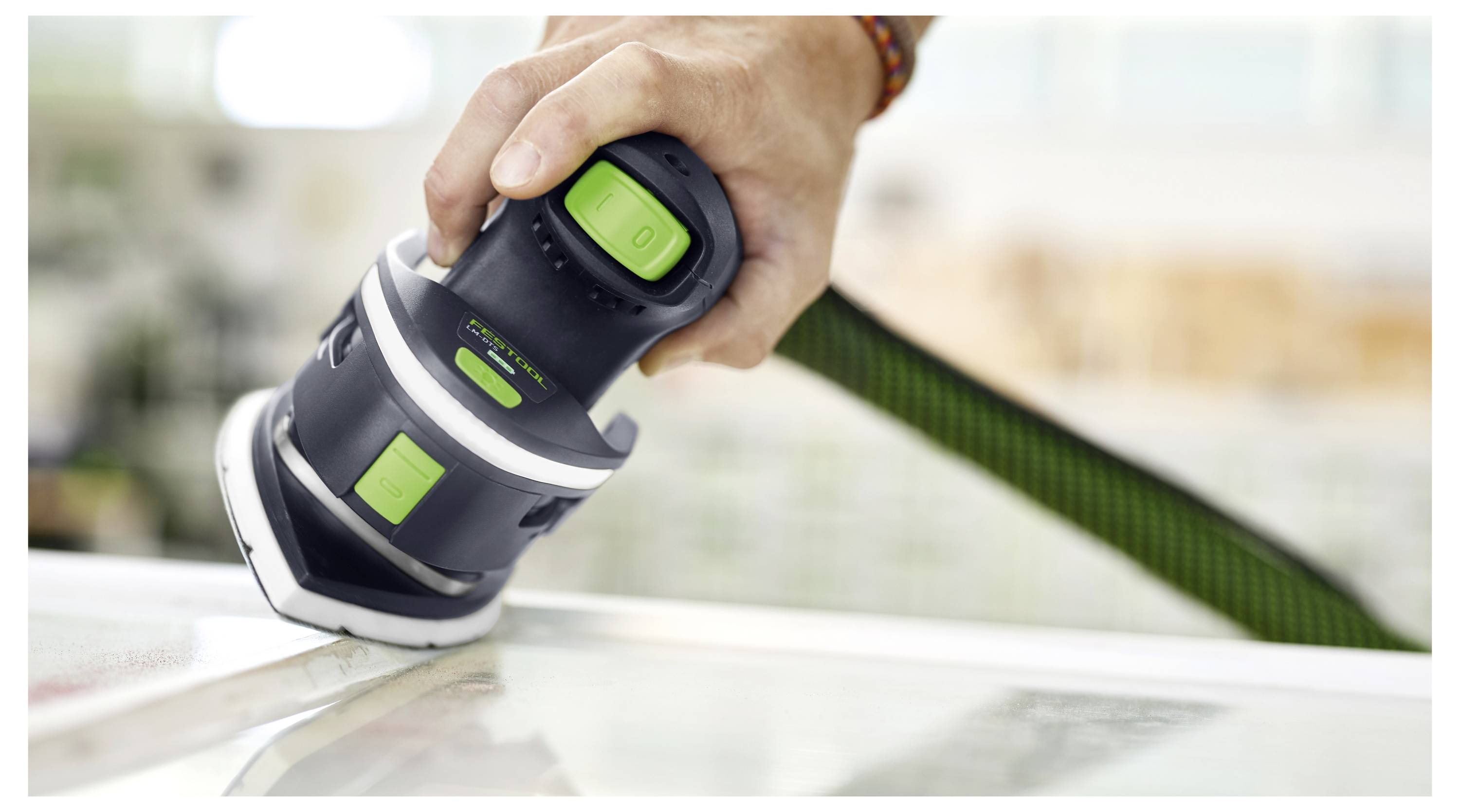 Close-up of a person using a handheld electric sander on a wooden surface. The sander is black with green buttons and features.
