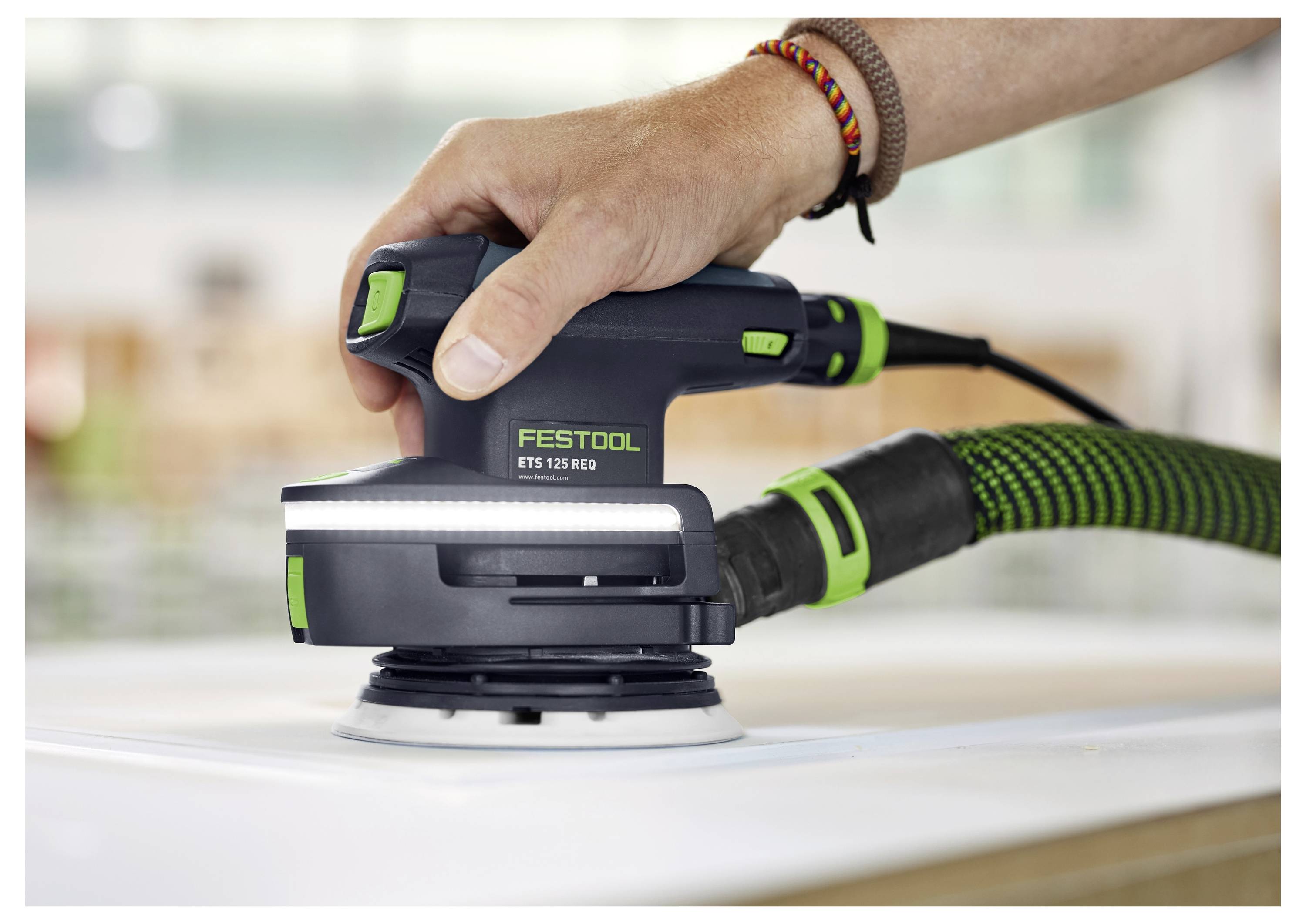 Une main actionnant une ponceuse Festool ETS 125 REQ sur une surface plane, connectée à un tuyau d'aspiration, démontrant ses capacités de ponçage.