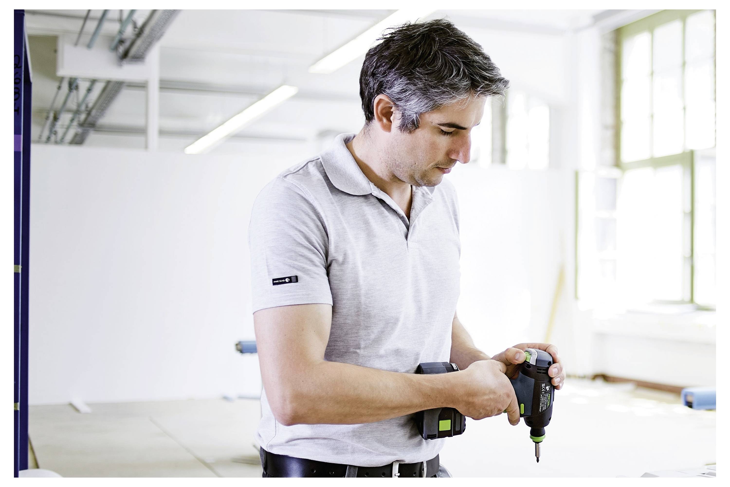 A man in a light gray polo shirt is using a power drill inside a bright, spacious workshop or studio. The environment appears industrial with windows and white walls.
