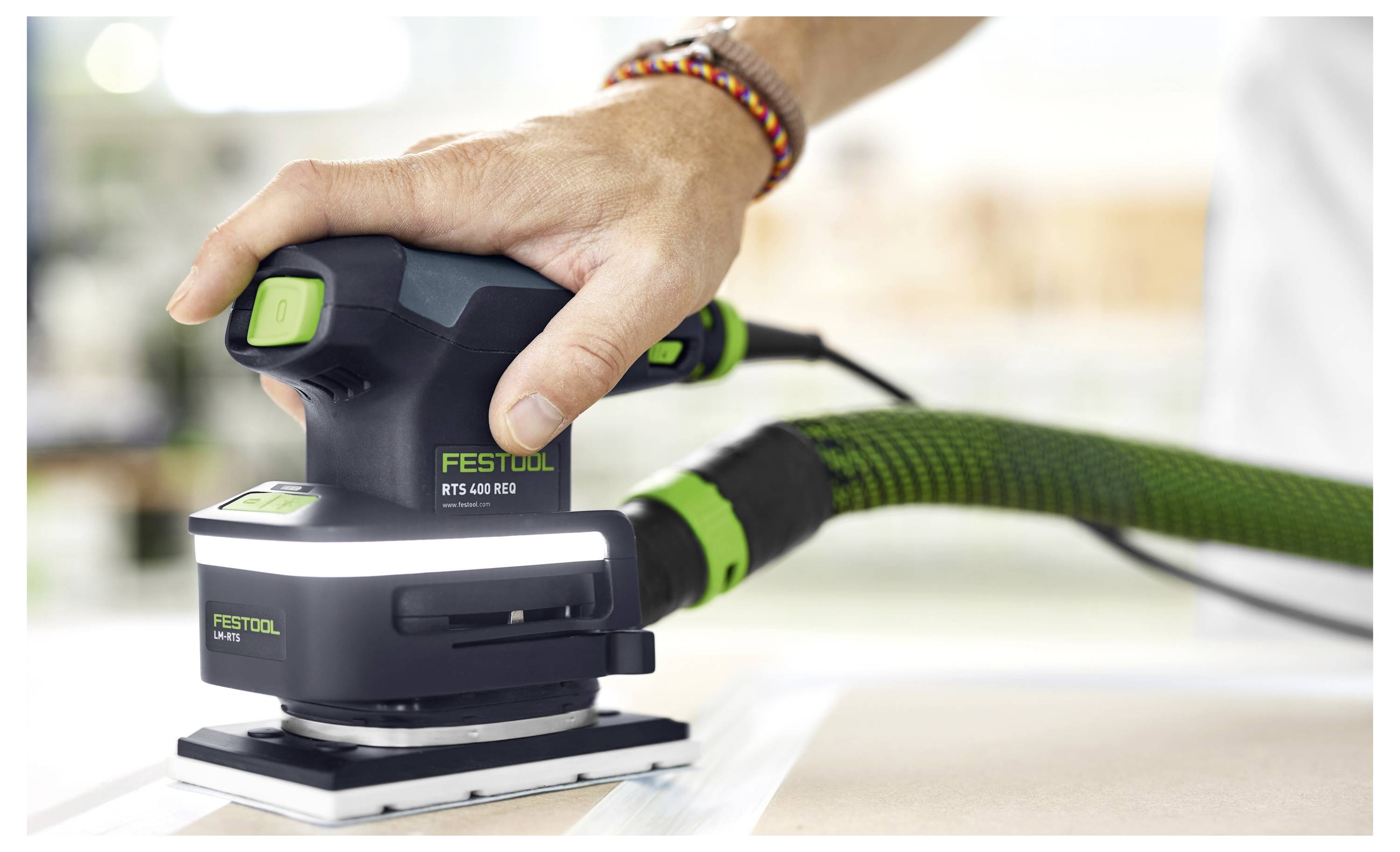 Une personne utilise une ponceuse Festool RTS 400 REQ sur une surface plane avec un tuyau d'aspirateur connecté, en mettant l'accent sur une extraction efficace de la poussière.