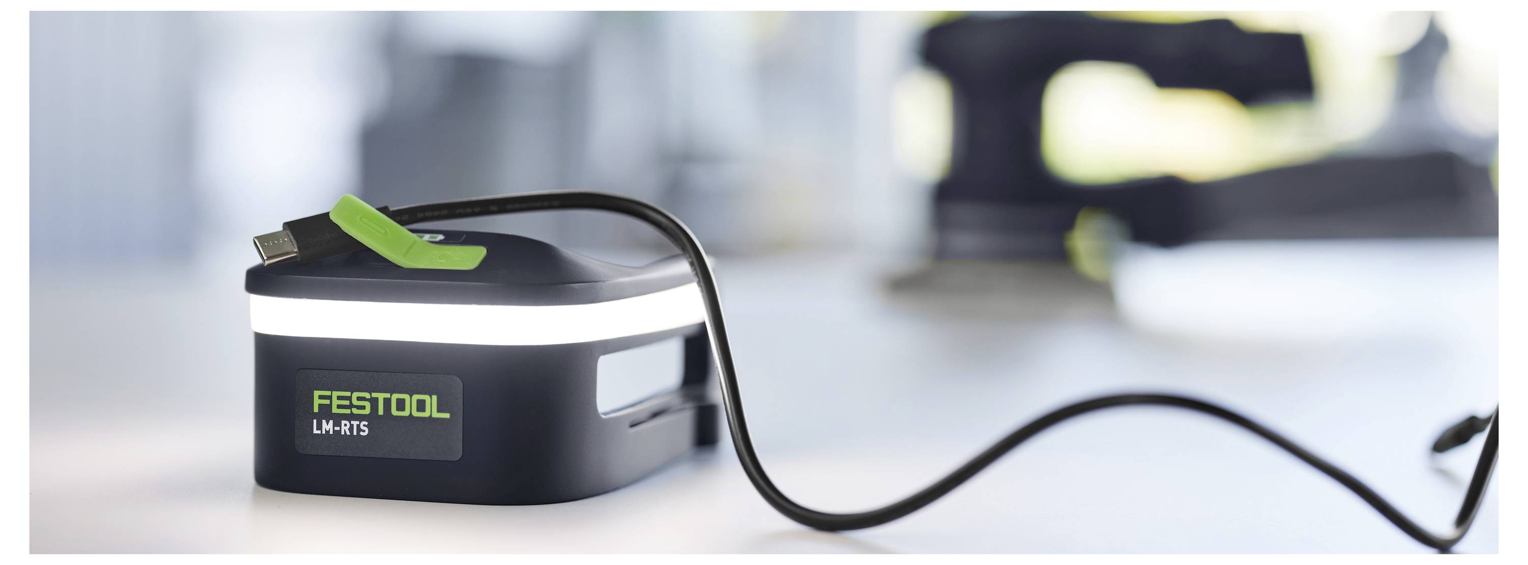 « Un bloc-batterie Festool avec un câble de charge sur une surface de couleur claire, arrière-plan d'atelier avec outils partiellement flou. »